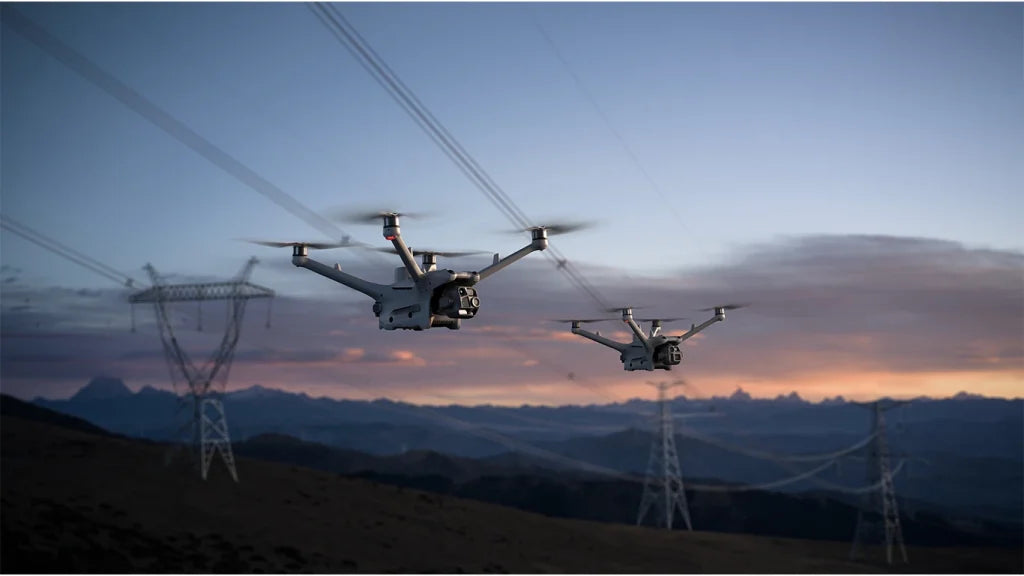 Zwei Matrice 4D und Matrice 4TD Drohnen fliegen in der Abenddämmerung in der Nähe von Stromleitungen, im Hintergrund sind Berge und ein bewölkter Himmel zu sehen. Die Drohnen scheinen die elektrische Infrastruktur zu inspizieren oder zu überwachen.