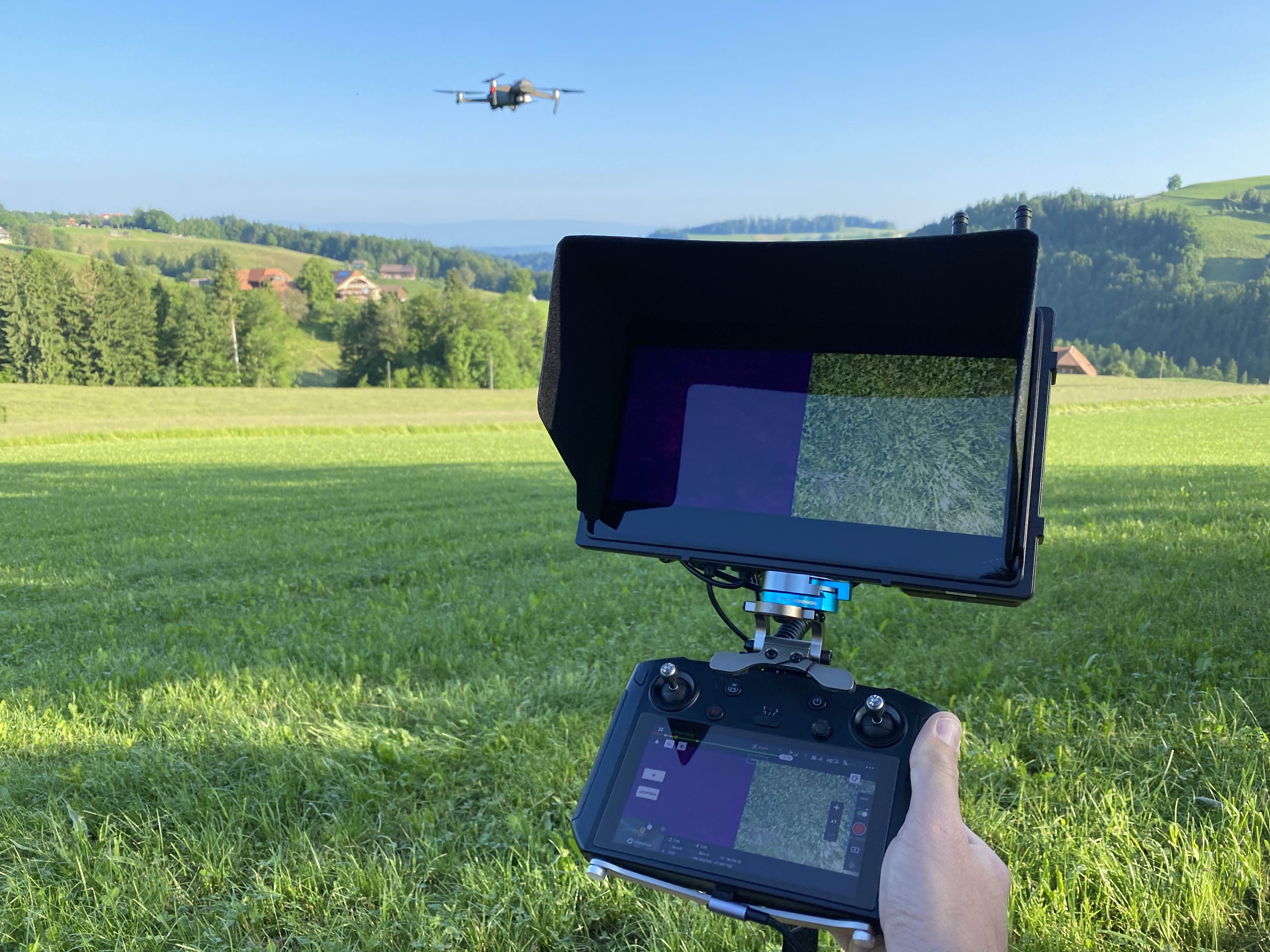 Die Hand einer Person greift eine Drohnenfernbedienung mit integriertem Bildschirm, ähnlich einer Monitorhalterung. Auf diesem werden Grasfelder angezeigt, während die Drohne über Hügel und Bäume schwebt. Die nahtlose Steuerung wird durch das Kondor Blue Mini Schnellverschlusssystem gewährleistet.
