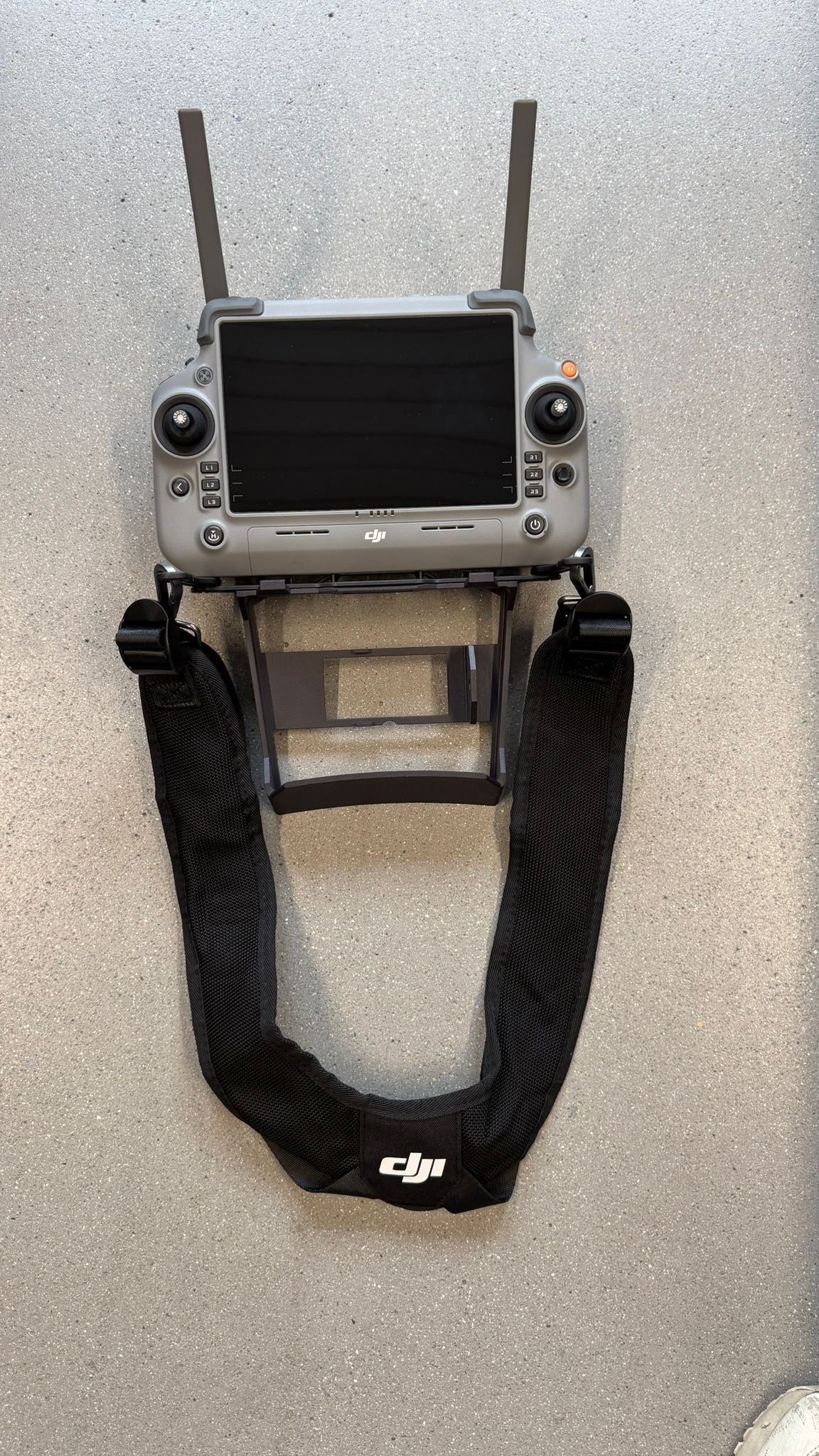 Eine CIELOTEC RC Plus 2-Distanzhalterung – SDR- & Lautsprecher-Version (3D-Druck PA12) ist an einer DJI RC Plus-Fernbedienung mit Display und zwei Antennen befestigt, die wiederum an einem schwarzen DJI-Tragegurt mit DJI-Logo angebracht ist. Die gesamte Anlage, einschließlich des 3D-gedruckten PA12-Bauteils, ruht sicher auf einer grauen Oberfläche.