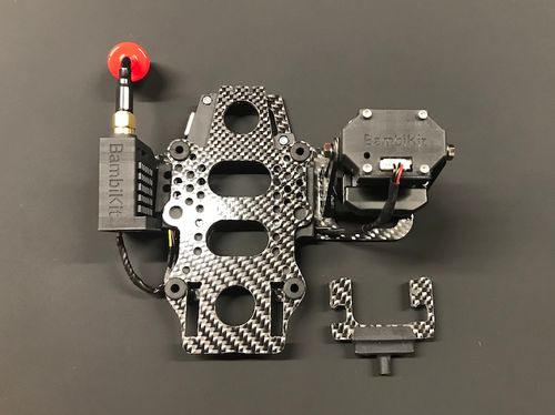 Ein schwarzer FPV-Drohnenrahmen aus Kohlefaser mit angebrachten elektronischen Komponenten und einer Antenne, positioniert auf einer dunklen Oberfläche, mit einem separaten Montageteil daneben.