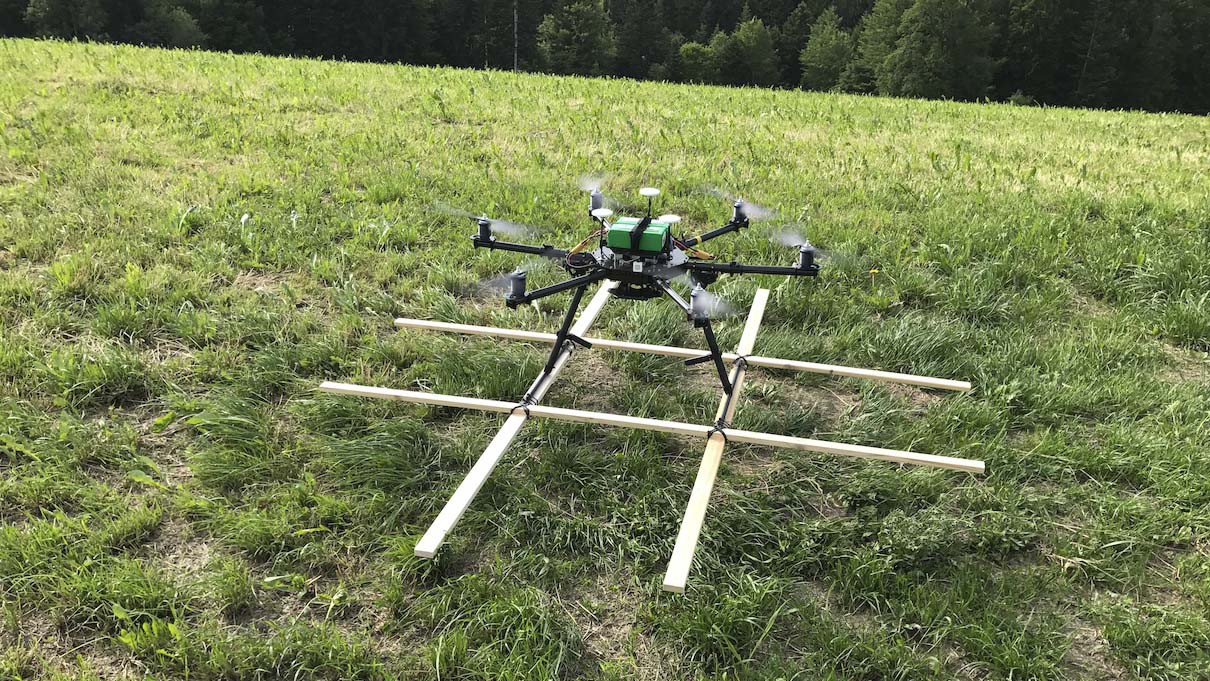Eine Drohne mit einer grünen Zentraleinheit und mehreren Rotoren schwebt über einer Wiese und trägt einen rechteckigen Holzrahmen unter sich. Im Hintergrund sind Bäume zu sehen.