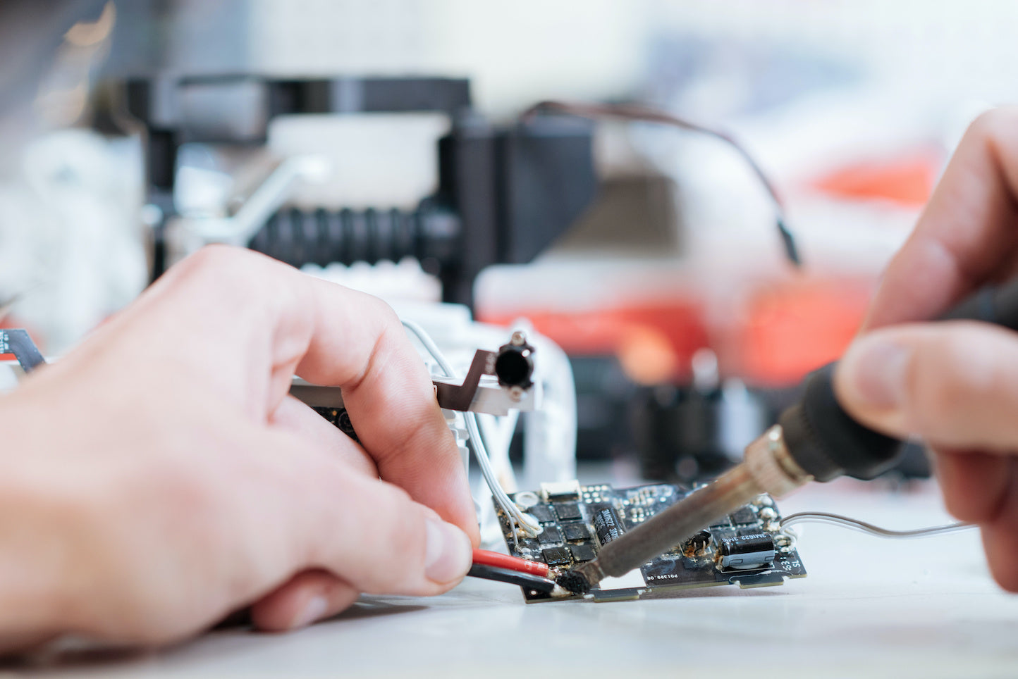 Nahaufnahme von Händen, die mit einem Lötkolben Drähte auf eine Leiterplatte löten, umgeben von elektronischen Bauteilen und unscharfer Ausrüstung im Hintergrund.