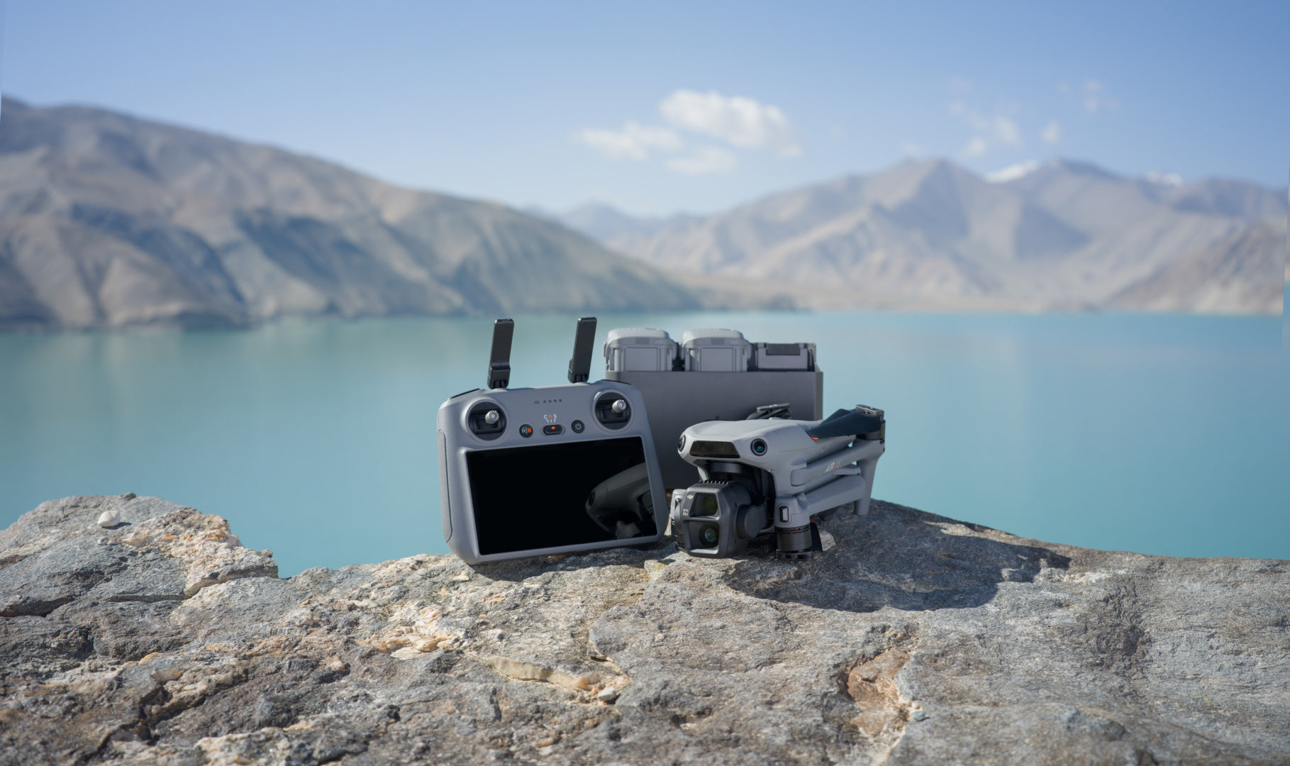 Eine DJI Air 3S Fly More Combo (RC 2) von DJI Consumer mit einem 1-Zoll-CMOS-Sensor und Dual-Kamerasystem steht auf einer felsigen Oberfläche mit Blick auf einen ruhigen türkisfarbenen See, der von dunstigen Berggipfeln unter einem klaren blauen Himmel eingerahmt wird – eine idyllische Landschaft für atemberaubende Aufnahmen.