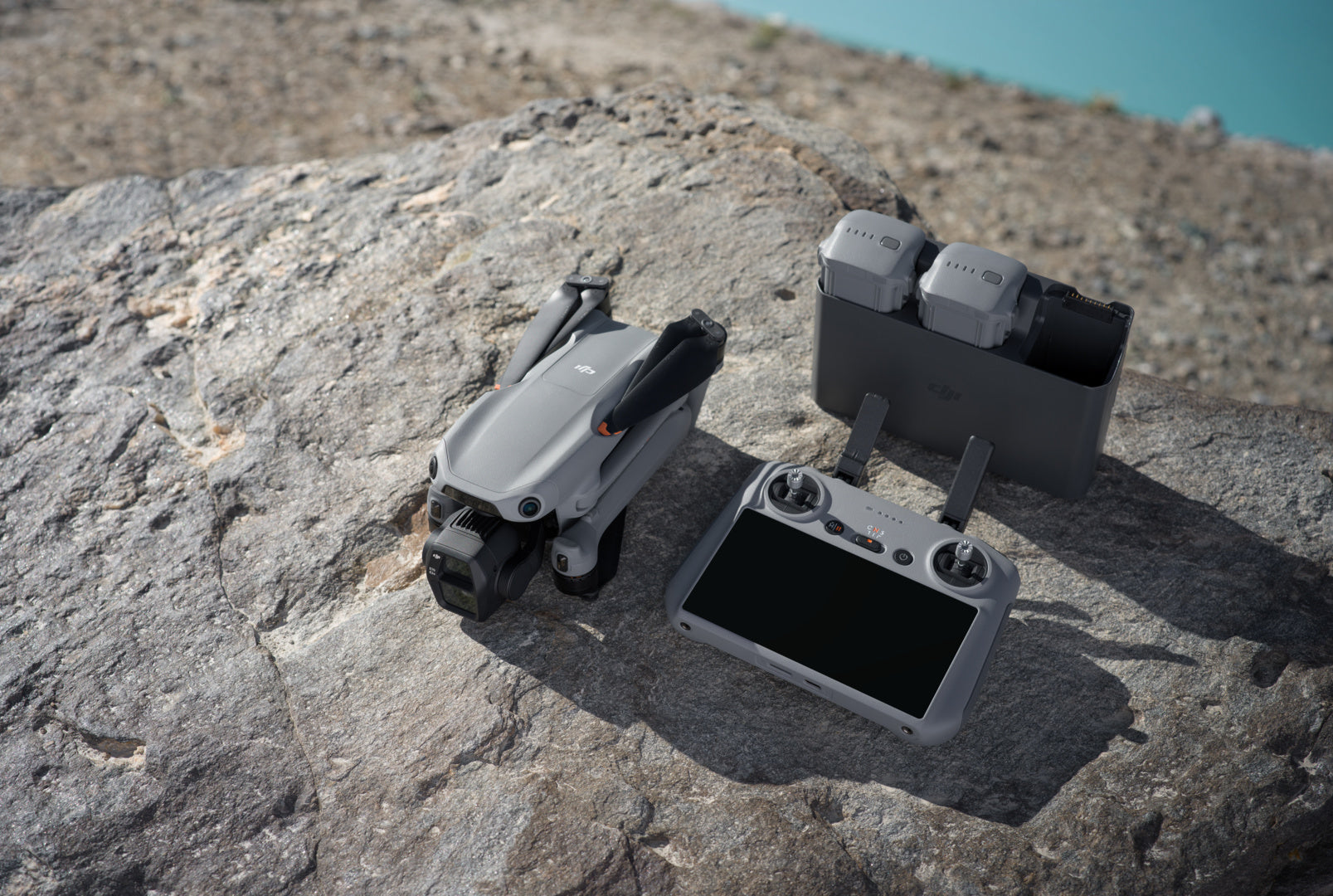 Eine DJI Air 3S Fly More Combo (RC 2) liegt zusammengeklappt auf einem großen Felsen. Sie ist mit einem Dual-Kamera-System und einer Fernbedienung mit Bildschirm ausgestattet. In der Nähe liegen zwei Akkus in der Ladestation vor einem kiesigen Hintergrund, der in strahlend blaues Wasser mündet.