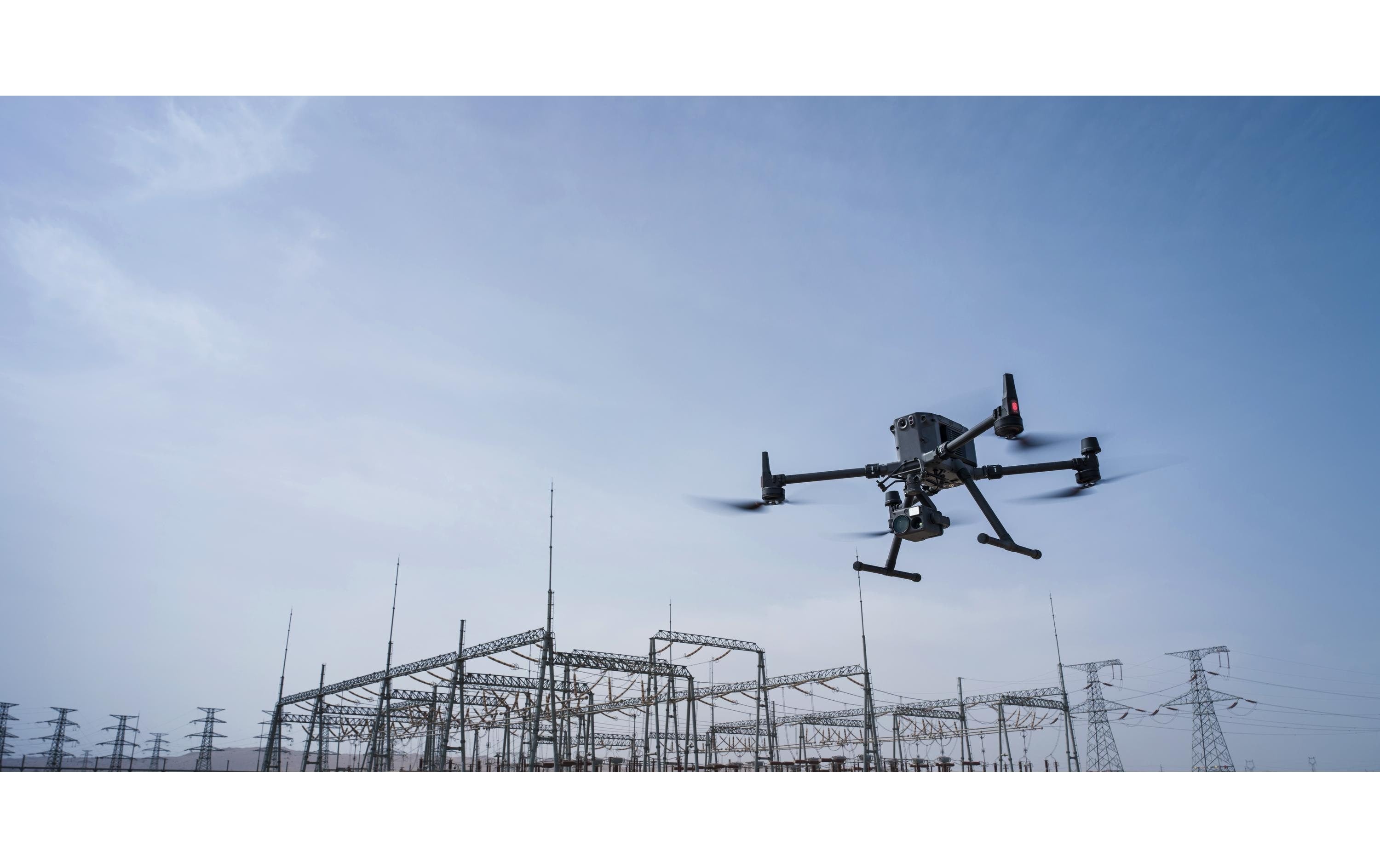 Ein DJI Enterprise Multikopter Matrice 350 RTK (EU) DJI Care Basic fliegt über eine elektrische Umspannstation mit Stromleitungen und Türmen unter blauem Himmel.