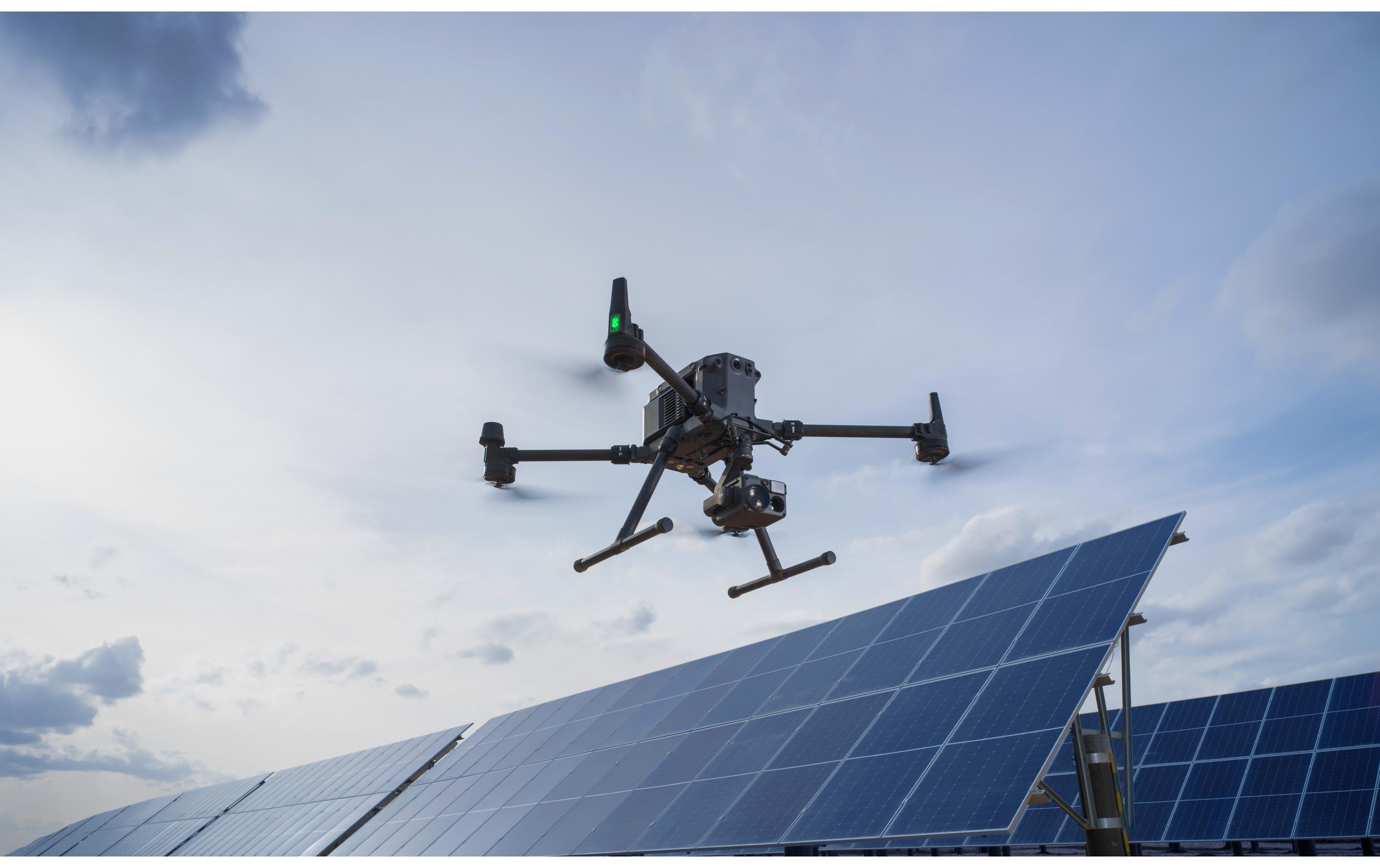 Ein DJI Enterprise Multikopter Matrice 350 RTK (EU) mit DJI Care Basic von DJI Enterprise schwebt über Solarpanelen unter teils bewölktem Himmel, vermutlich zur Inspektion oder für Luftaufnahmen der Anlage.