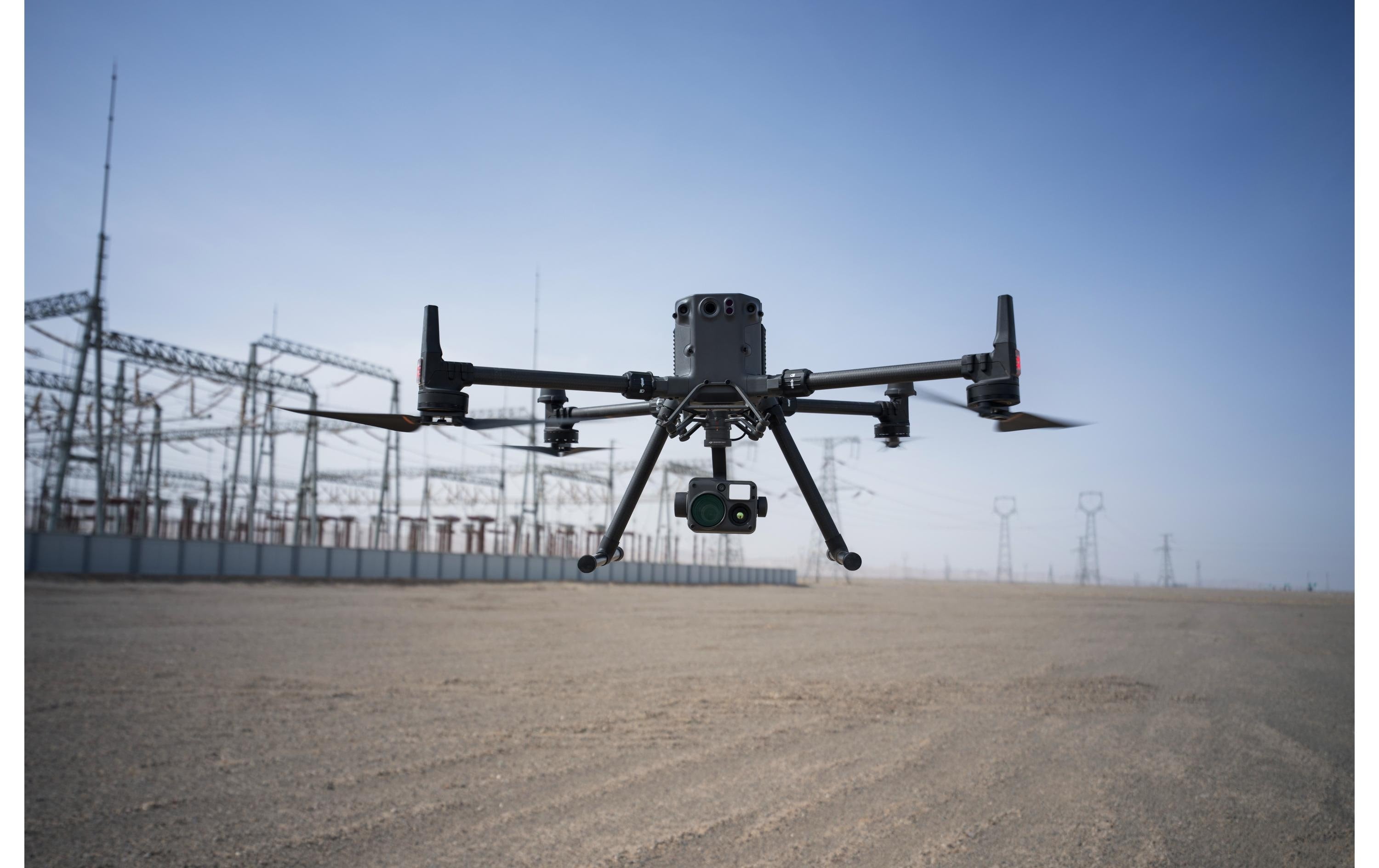 Der DJI Enterprise Multikopter Matrice 350 RTK (EU) DJI Care Basic schwebt für Luftaufnahmen knapp über dem Boden nahe eines Industriegeländes mit Strommasten und Hochspannungsleitungen unter blauem Himmel.