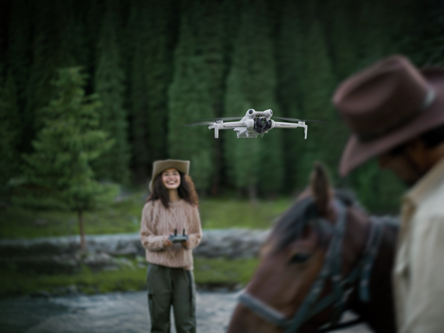 Eine Person mit Hut bedient eine DJI Mini 4 Pro Fly More Combo (RC 2) in der Nähe eines Waldes und lächelt neben einer Person zu Pferd. Üppige grüne Bäume und ein kleiner Bach dienen als Hintergrund, während die Drohne ihre Bildgebungsleistung demonstriert.
