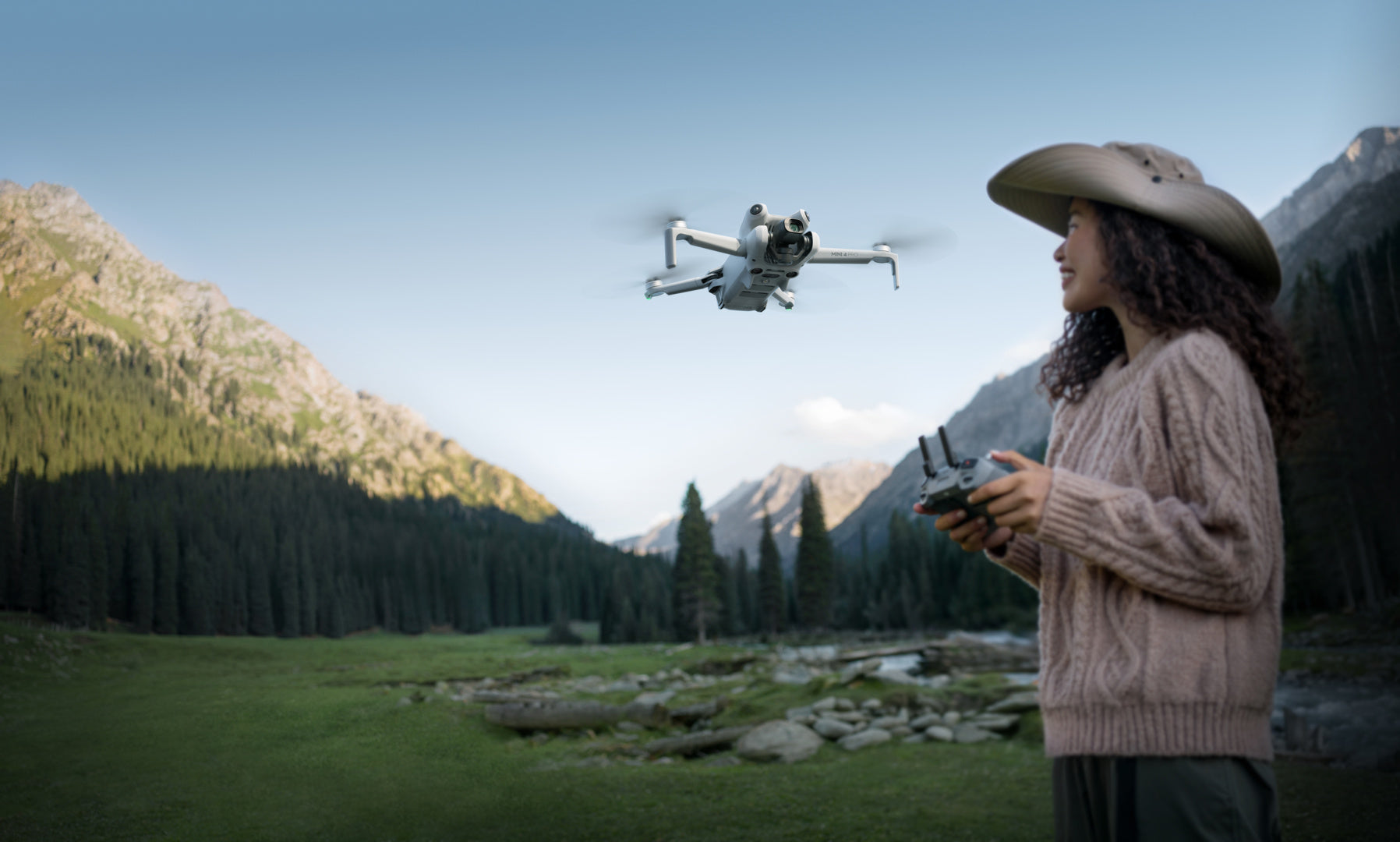 Mit breitkrempigem Hut und Pullover bekleidet, steuert eine Person gekonnt eine DJI Mini 4 Pro Fly More Combo (RC 2) von DJI Consumer. Die Hindernisvermeidungsfunktion der Drohne sorgt für einen ruhigen Flug über ein Tal mit immergrünen Bäumen und Bergen unter klarem Himmel.