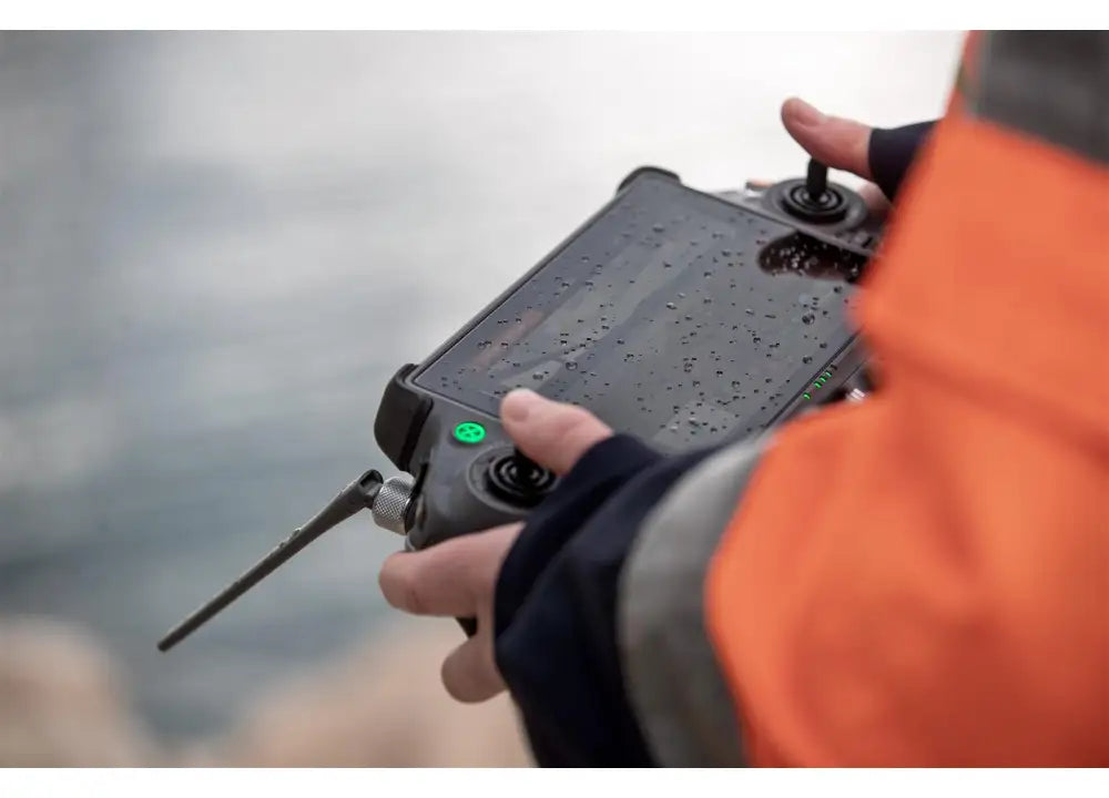 Eine Person in einer orangefarbenen Jacke hält eine schwarze DJI Enterprise RC Plus 2 Enterprise-Fernsteuerung (M4 Serie) mit Regentropfen darauf. Ihr leuchtstarkes Display sorgt für klare Sicht auch bei Regen, da sie im Freien vor einem wässrigen Hintergrund steht.