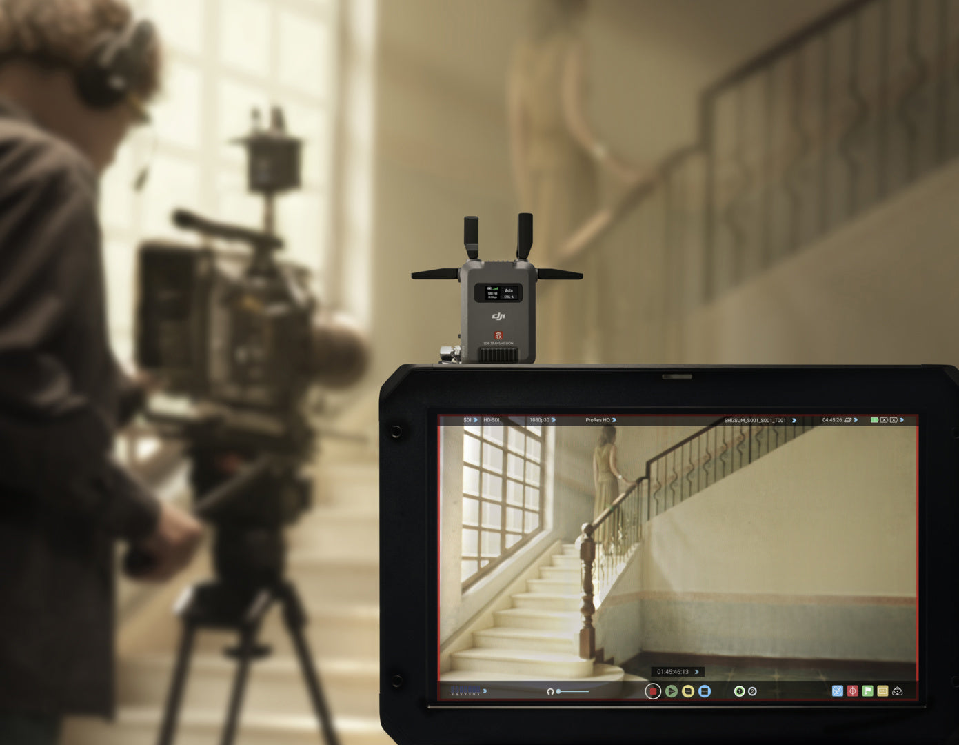 Ein Kameramann filmt mit der DJI Pro Digitale Bildübertragung – DJI SDR Transmission Combo (Reichweite bis zu 3 km) eine Treppenszene und zeigt Live-Aufnahmen auf einem Monitor in einer eleganten, gut beleuchteten Umgebung mit großen Fenstern und verziertem Geländer.