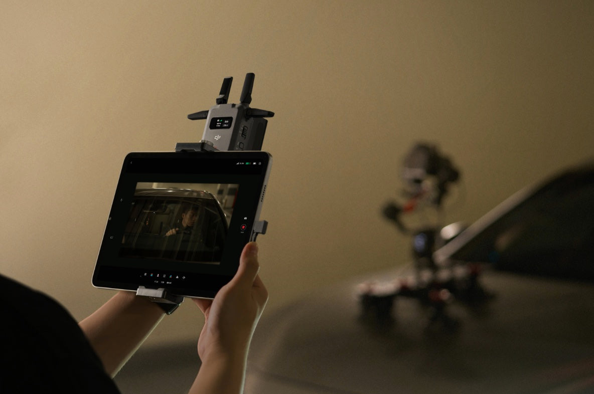 Eine Person hält ein Tablet, auf dem eine Videoszene gezeigt wird. Im Hintergrund ist ein Kamera-Rig auf einer Motorhaube zu sehen. Das Setup nutzt die DJI Pro Digitale Bildübertragung – DJI SDR Transmission Combo und ermöglicht Filmproduktionen mit einer Reichweite von bis zu 3 km.