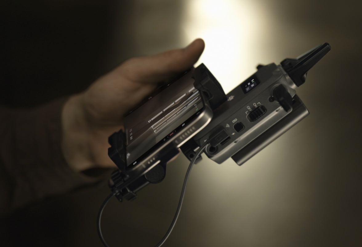 Eine Nahaufnahme zeigt eine Hand, die die DJI Pro Digitale Bildübertragung – DJI SDR Transmission Combo hält, verbunden mit einem Smartphone. Das Gerät verfügt über Drehregler, Schalter, Display und eine Reichweite von bis zu 3 km mit eingesteckten Kabeln.