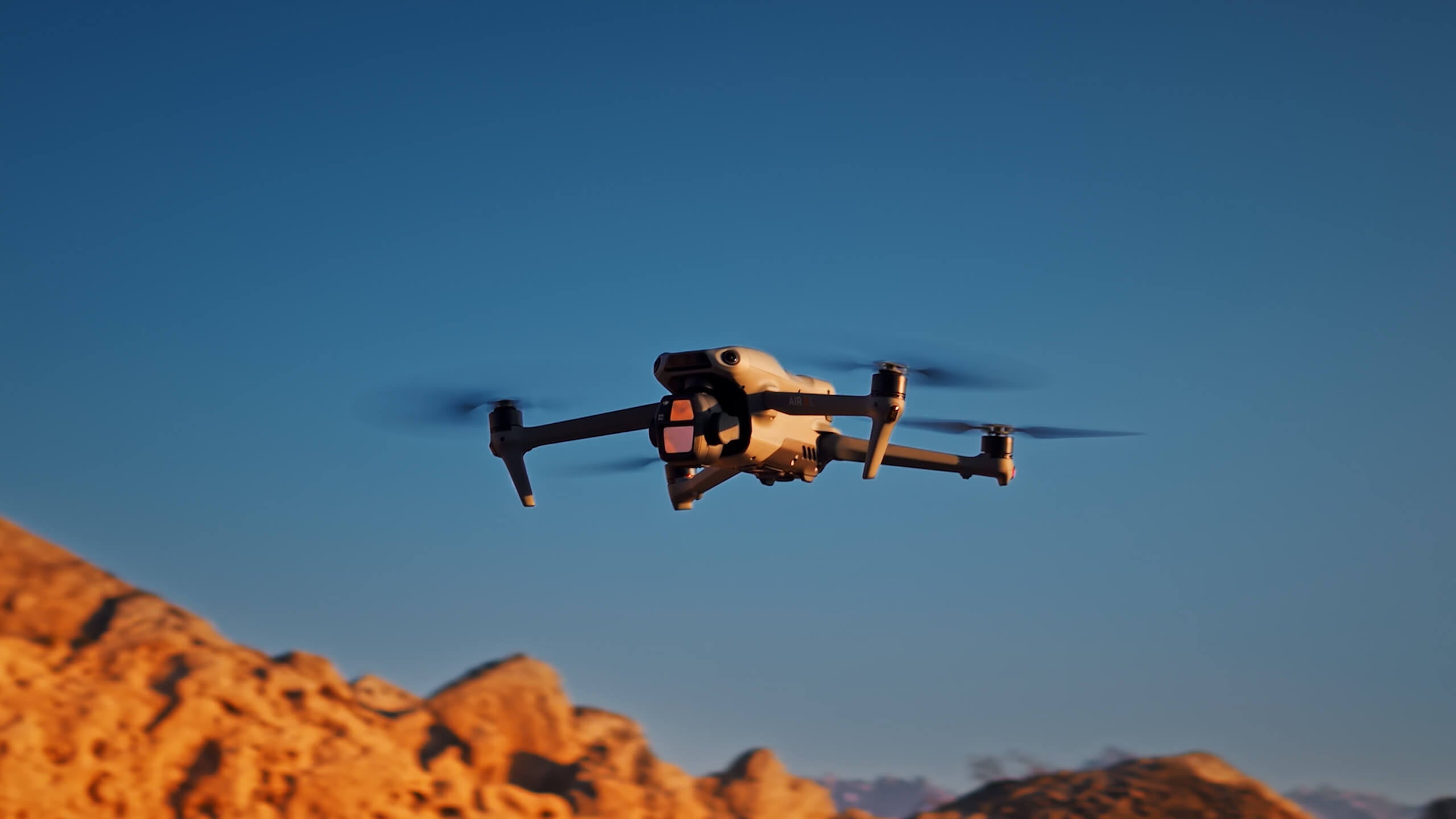 Die DJI Air 3S Fly More Combo (RC 2) von DJI Consumer fliegt vor blauem Himmel und nimmt mit ihrem Dual-Kamera-System und dem 1-Zoll-CMOS-Sensor atemberaubende Bilder auf. Darunter unterstreicht das sonnige Wüstengelände die Schönheit der Natur.