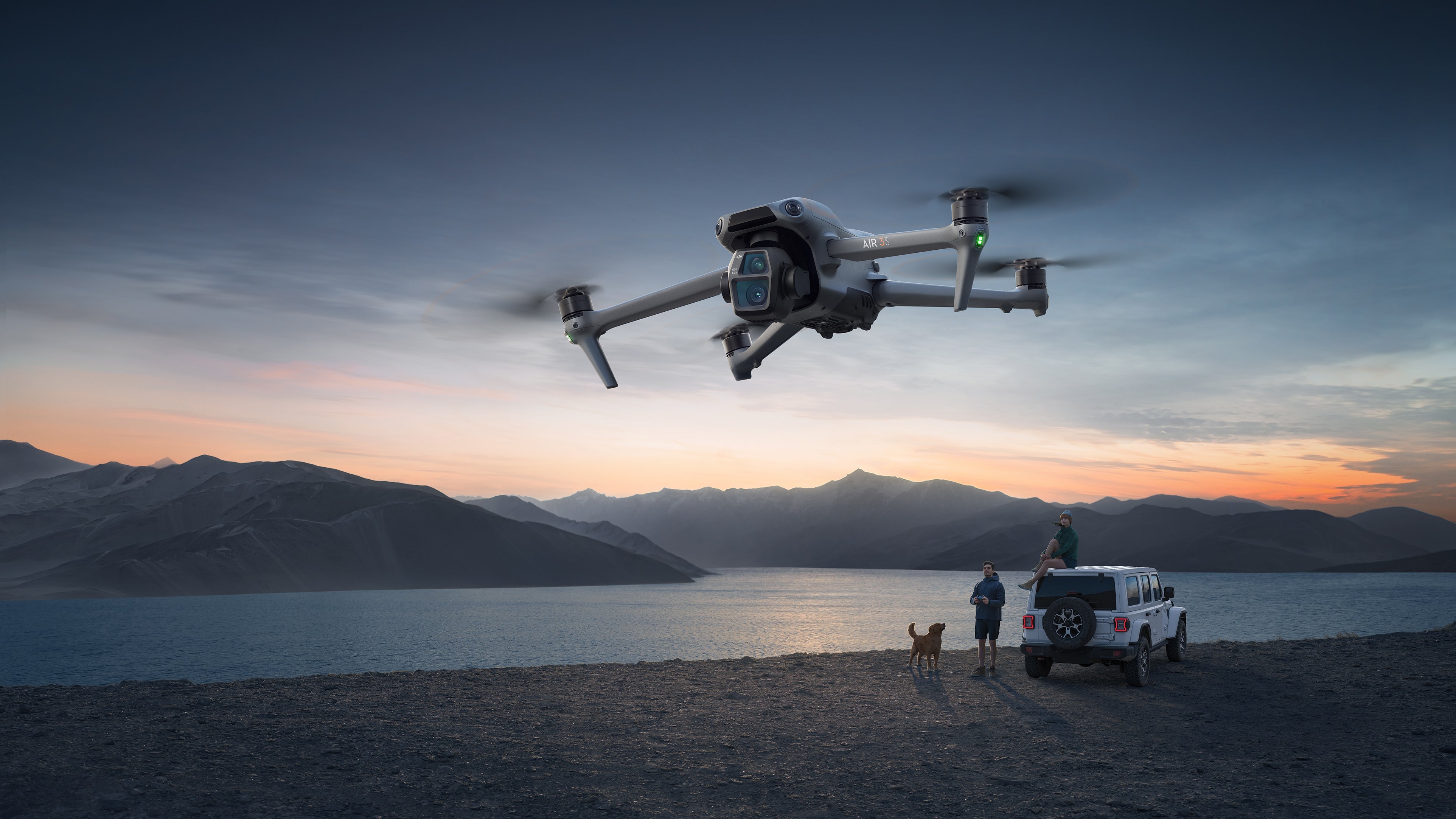 Die DJI Air 3S Fly More Combo (RC 2) von DJI Consumer, ausgestattet mit einem Dual-Kamerasystem und einem 1 Zoll CMOS-Sensor, schwebt bei Sonnenuntergang über einem malerischen Seeufer. Darunter bewundern zwei Personen in einem Jeep mit einem Hund an der Seite die Bergsilhouetten, während der Himmel von Orange zu Blau wechselt.