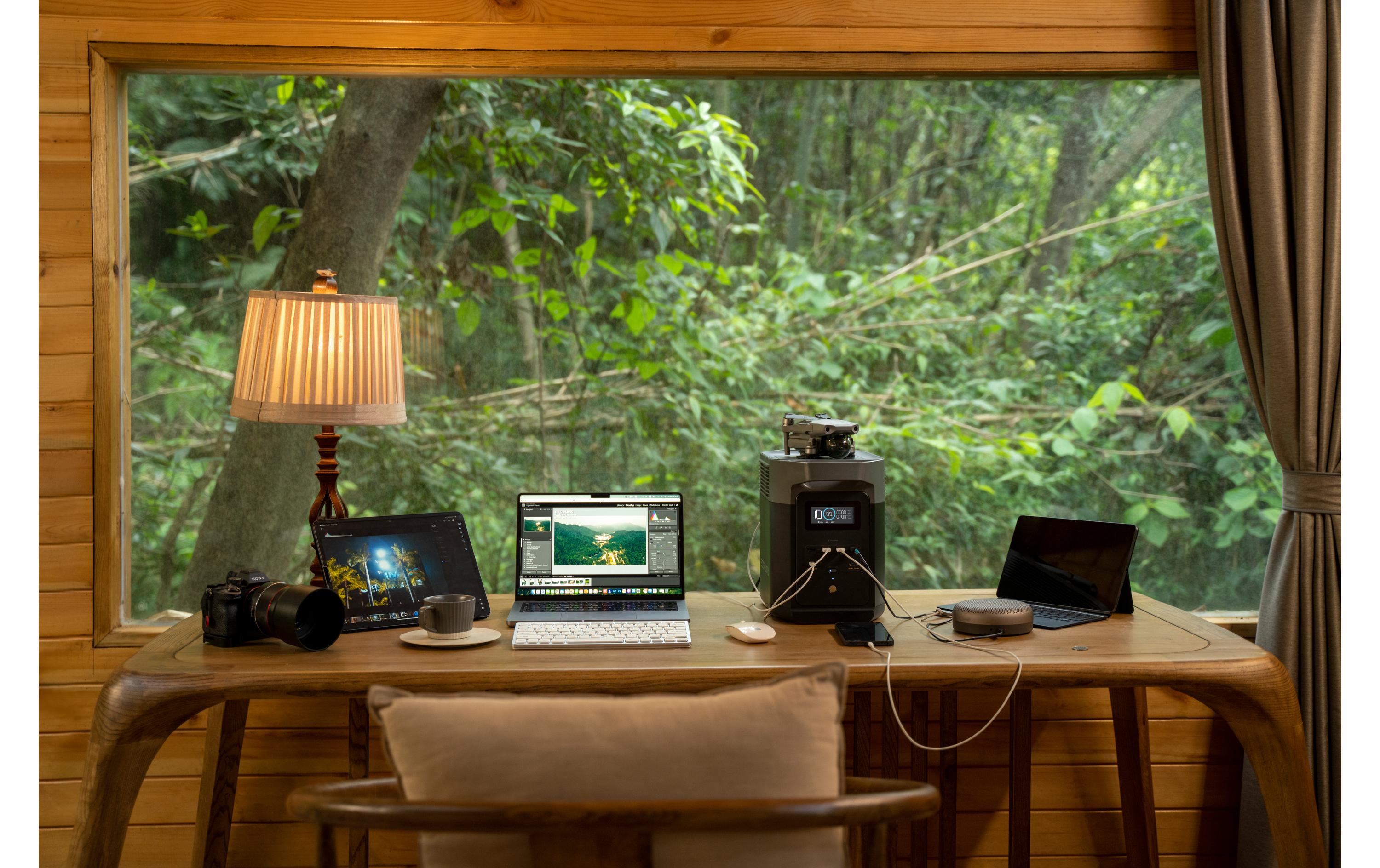 Ein gemütlicher Arbeitsbereich mit einem Holzschreibtisch an einem großen Fenster mit Blick auf einen üppigen Wald ist mit Laptop, Tablet, Kamera und Kaffeemaschine ausgestattet – alles nahtlos mit Strom versorgt von der EcoFlow Power Station Delta 2, 1000 Wh. Eine Tischlampe beleuchtet den gepolsterten Stuhl vor dem Schreibtisch.