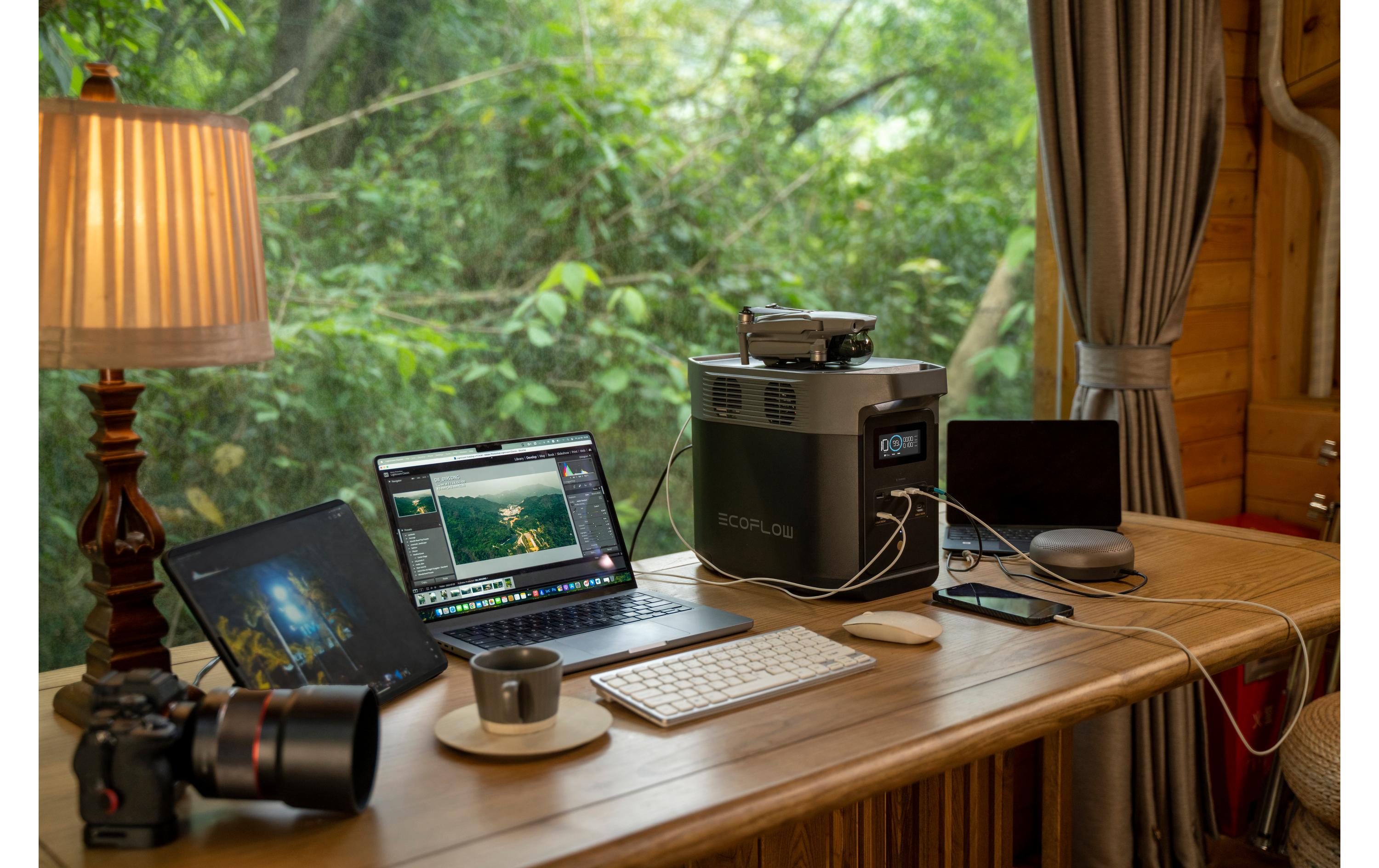 Ein gemütlicher Arbeitsplatz mit Laptop, Bearbeitungssoftware, Kamera und Tablet auf einem Holzschreibtisch. Eine Kaffeetasse steht neben einer EcoFlow Power Station Delta 2 (1000 Wh). Sie wird von einer Lampe am Fenster beleuchtet. Die EcoFlow-App sorgt für Konnektivität, während draußen üppiges Grün gedeiht.