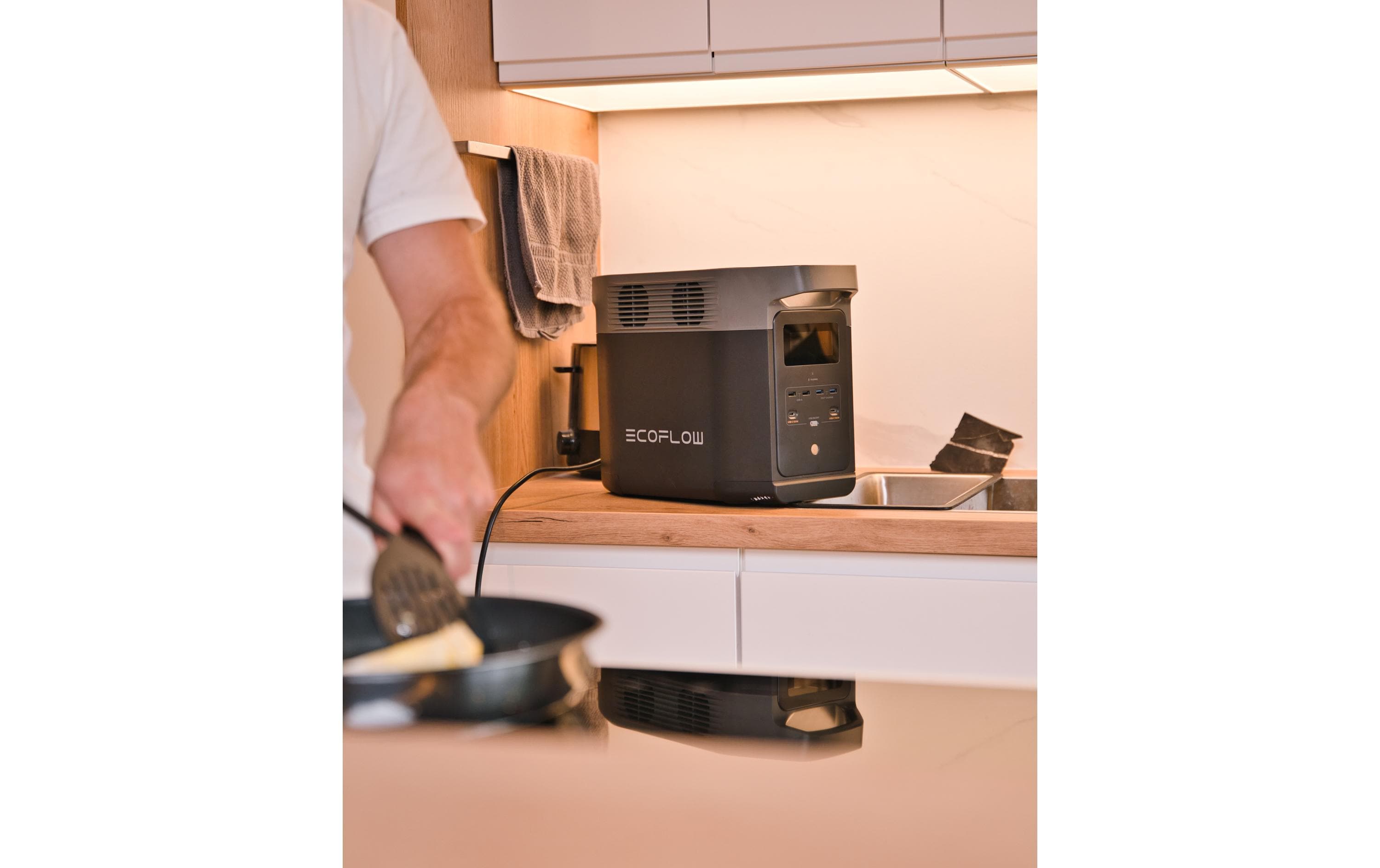 In einer modernen Küche kocht jemand an der Theke, neben ihm steht eine EcoFlow Power Station Delta 2, 1000 Wh. Mit der EcoFlow-App steuert er seine mobile Powerstation effizient, während Bratpfanne und Handtuch das schlichte Design betonen.