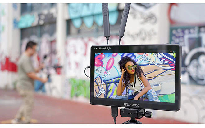 Der FEELWORLD Feelworld LUT11H 10,1-Zoll-HDMI-Monitor mit seinem ultrahellen 2000-Nit-Full-HD-Touchscreen-IPS-Display zeigt eine Frau mit Sonnenbrille und Trikot, die vor einer bunten Graffiti-Wand sitzt, während eine andere Person an einem lebendigen Wandgemälde im Hintergrund vorbeigeht.