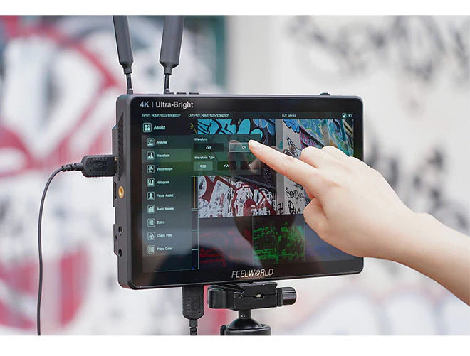 Eine Hand justiert die Einstellungen eines Feelworld LUT11H, eines 10,1-Zoll-FEELWORLD-Monitors mit ultrahellem 2000-Nit-Full-HD-Touchscreen-IPS-Display, der auf einem Stativ montiert ist. Der Bildschirm zeigt HDMI-Eingangsoptionen wie Wellenform und Histogramm vor lebendiger Graffiti-Kunst im Hintergrund.