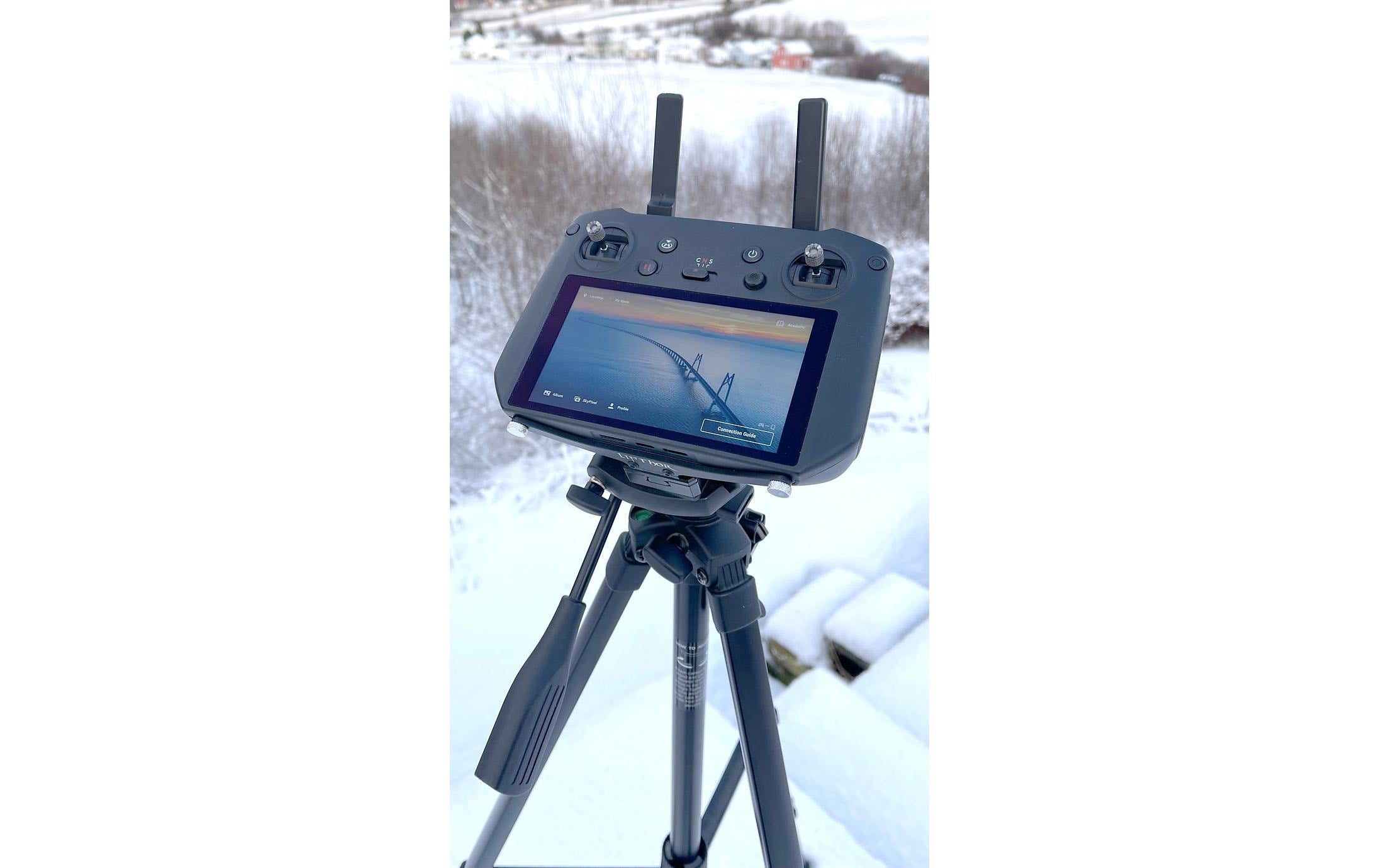 Ein DJI RC Pro Sender mit Bildschirm, montiert auf einer Lifthor-Stativhalterung für die Mavic 3 Serie, zeigt eine Luftaufnahme von verschneiten Bäumen und Boden. Zwei Antennen sind sichtbar.