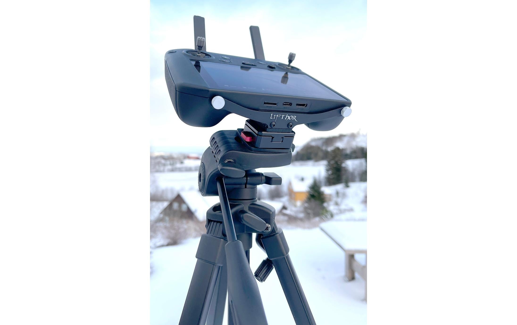 Eine LifThor-Stativhalterung dient zur Befestigung einer DJI RC Pro im Freien in einer verschneiten Landschaft. Der Controller verfügt über einen Bildschirm, Tasten und Antennen, die von oben herausragen. Im Hintergrund sind verschwommene Bäume und schneebedeckte Häuser zu sehen.