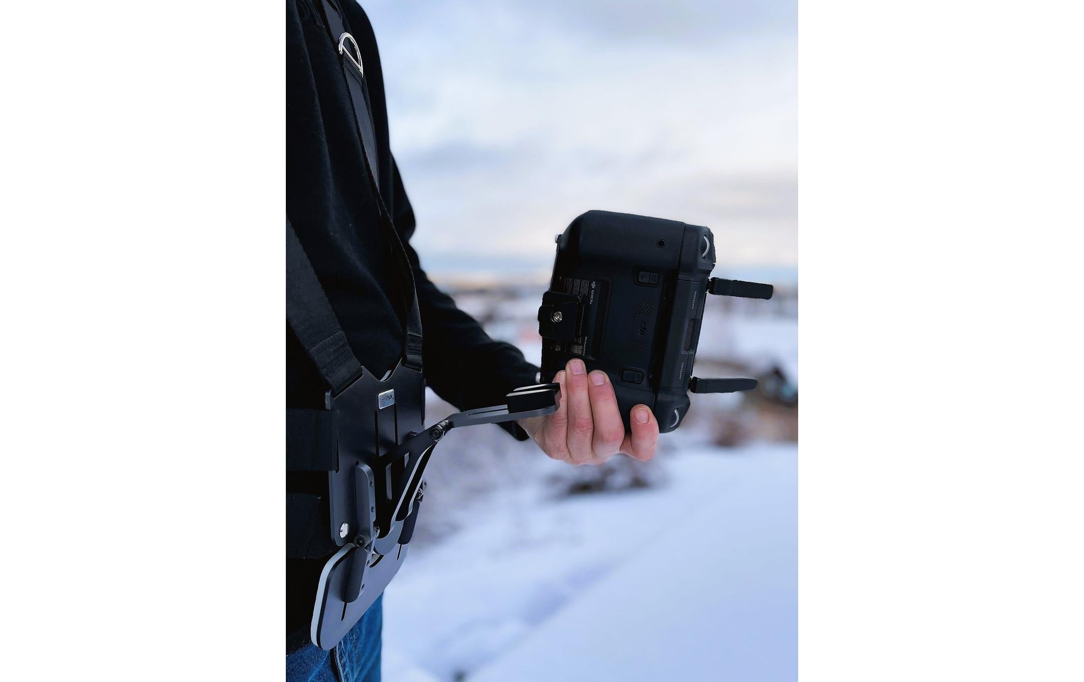 Draußen im Schnee verwendet eine Person fachmännisch die LifThor-Stativhalterung DJI RC Pro für die Mavic 3-Serie und hält sie mit einem Gurt fest, während sich darüber ein bewölkter Himmel abzeichnet.