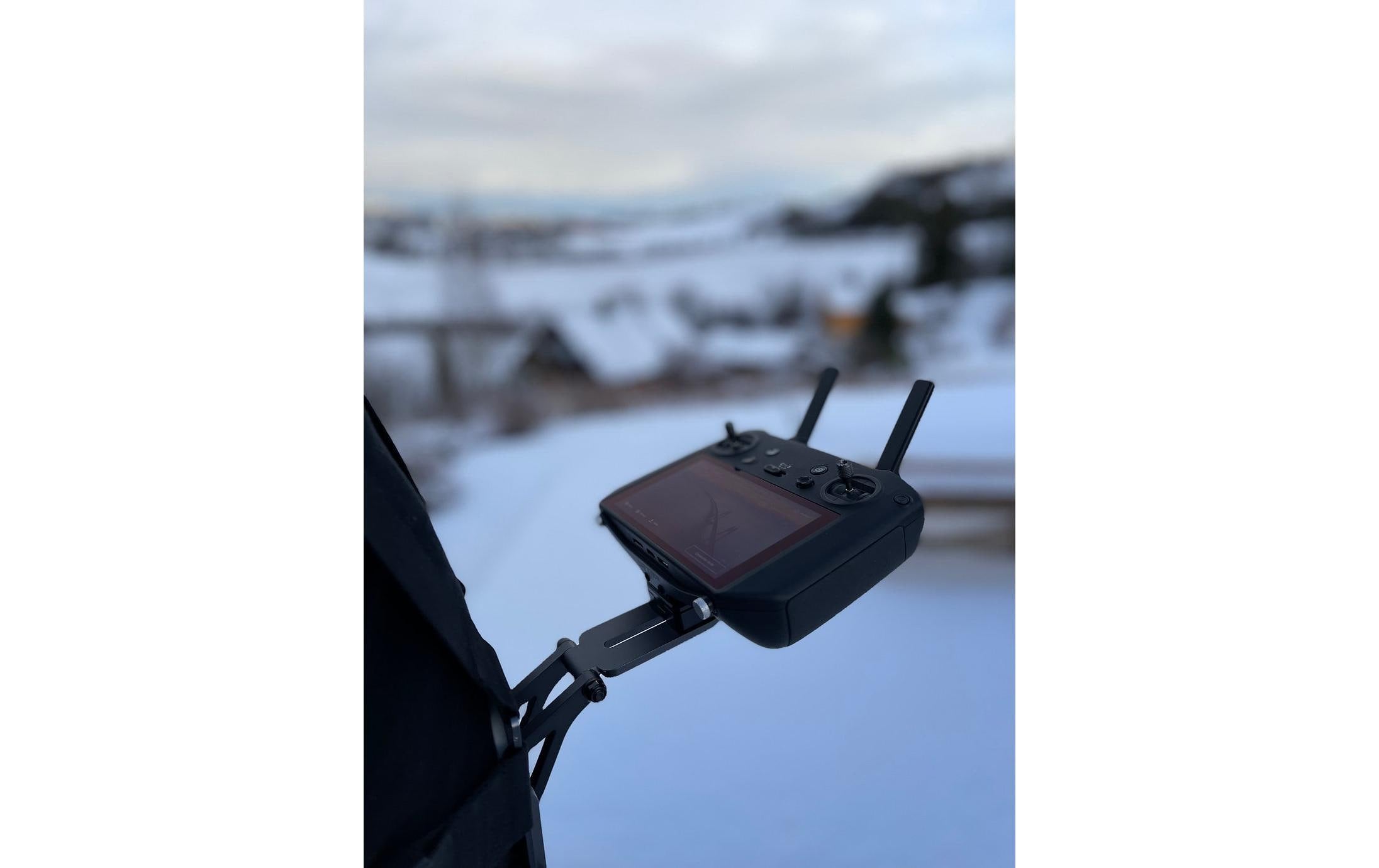 Eine Person hält die LifThor-Stativhalterung DJI RC Pro für die Mavic 3 Serie vor einer verschneiten Landschaft mit Dächern und Bäumen unter einem bewölkten Himmel.