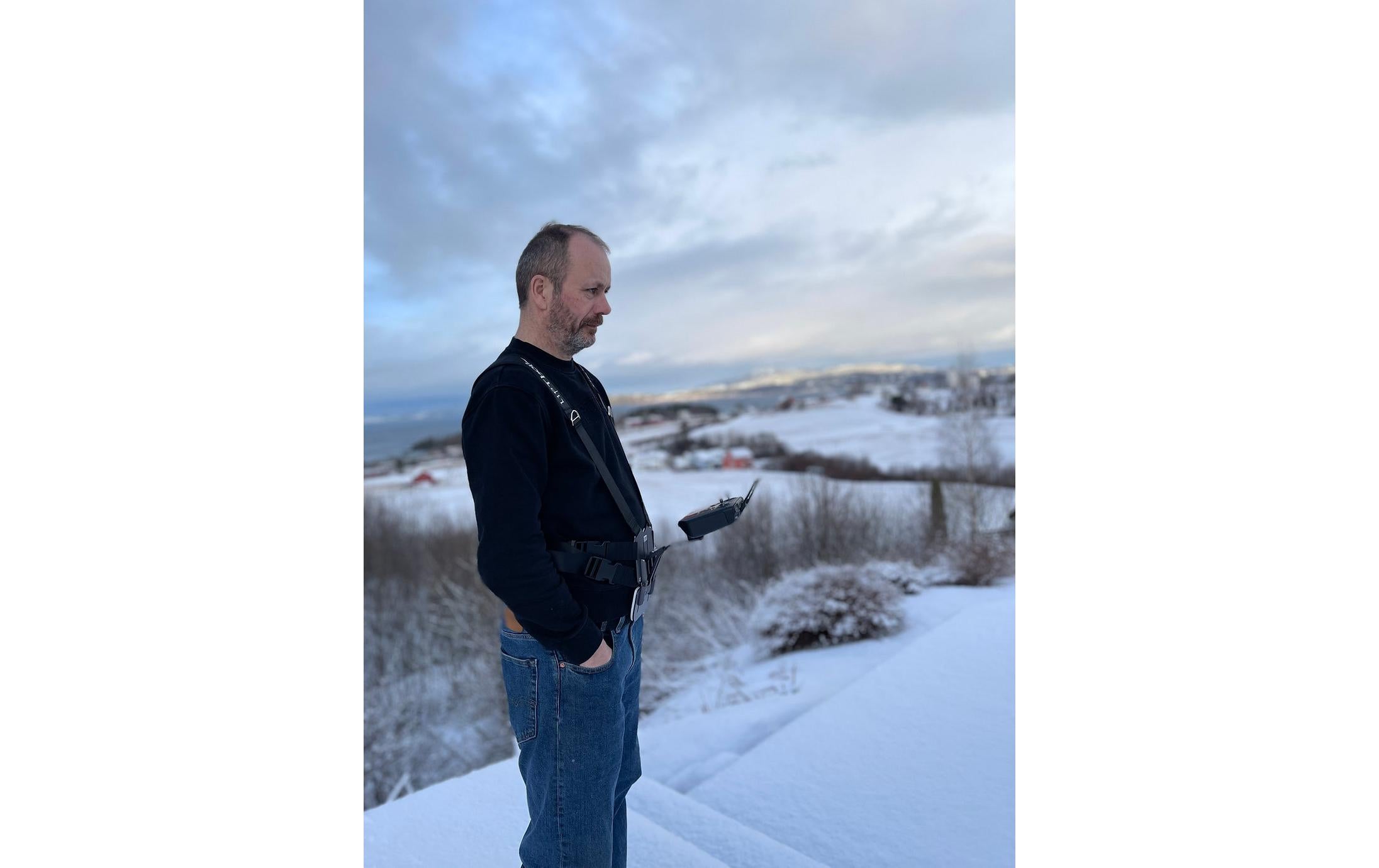 Ein Mann auf einem schneebedeckten Hügel konzentriert sich auf seine LifThor-Stativhalterung DJI RC Pro für die Mavic 3 Serie. Bekleidet mit einer dunklen Jacke und Jeans betrachtet er die Winterlandschaft mit fernen Bäumen und Häusern unter einem bewölkten Himmel, während er seine Mavic 3 Cine durch die frische Luft steuert.