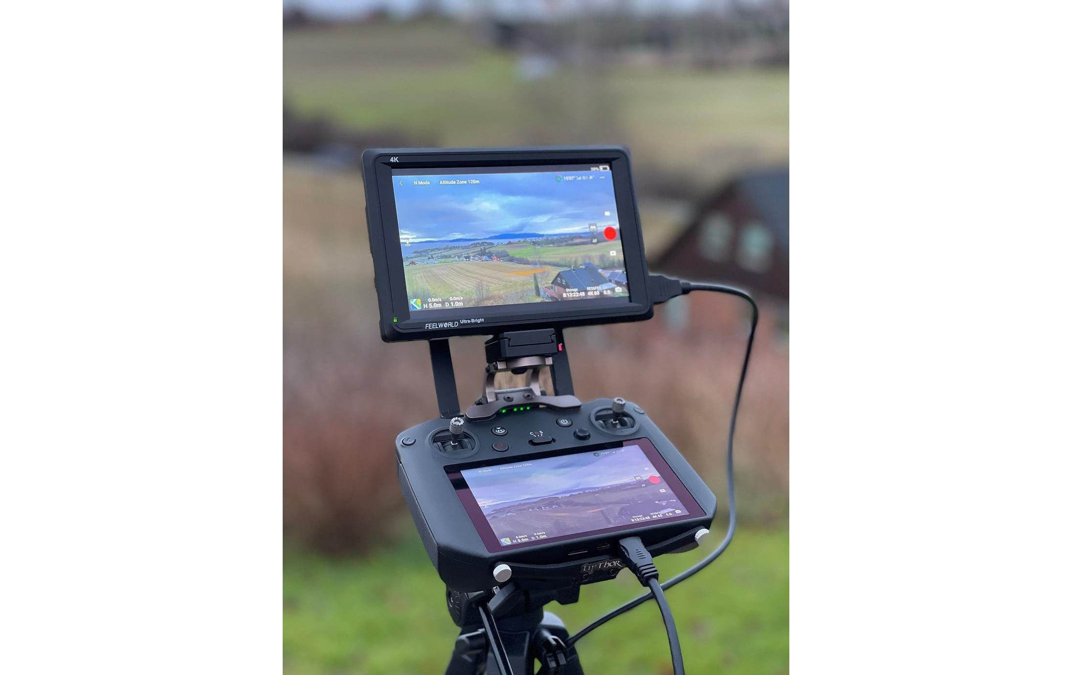 Nahaufnahme des LifThor Monitors & Stativhalterung II für DJI RC Pro. Zwei Bildschirme zeigen eine Live-Luftaufnahme einer ländlichen Landschaft. Das im Freien montierte Setup von Lifthor zeigt verschwommene Felder und ein Gebäude im Hintergrund.