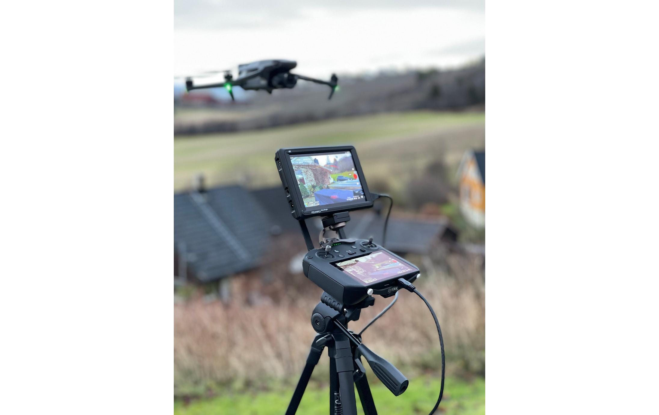 Ein LifThor Monitor & Stativ Halterung II für DJI RC Pro hält einen externen Bildschirm sicher fest, der den Live-Feed einer Drohne zeigt, die über einer Wiese schwebt und Häuser, Bäume und offene Felder unter einem bewölkten Himmel erfasst.