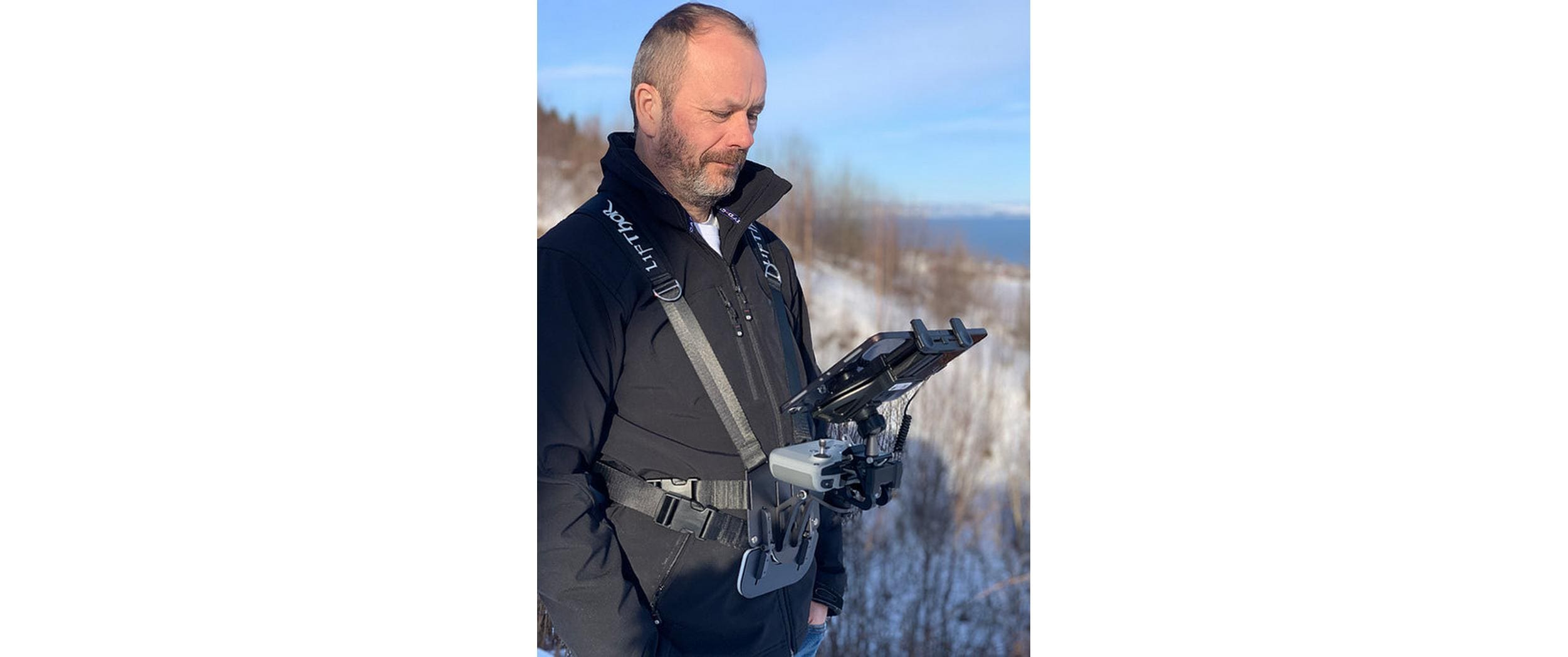 In einer verschneiten Landschaft nutzt ein Mann ein Tablet auf einem LifThor Sendertragegurt StrapThor PRO. Er trägt eine schwarze Jacke und konzentriert sich konzentriert auf den Bildschirm, vor der Kulisse von Bäumen und einem klaren Himmel.