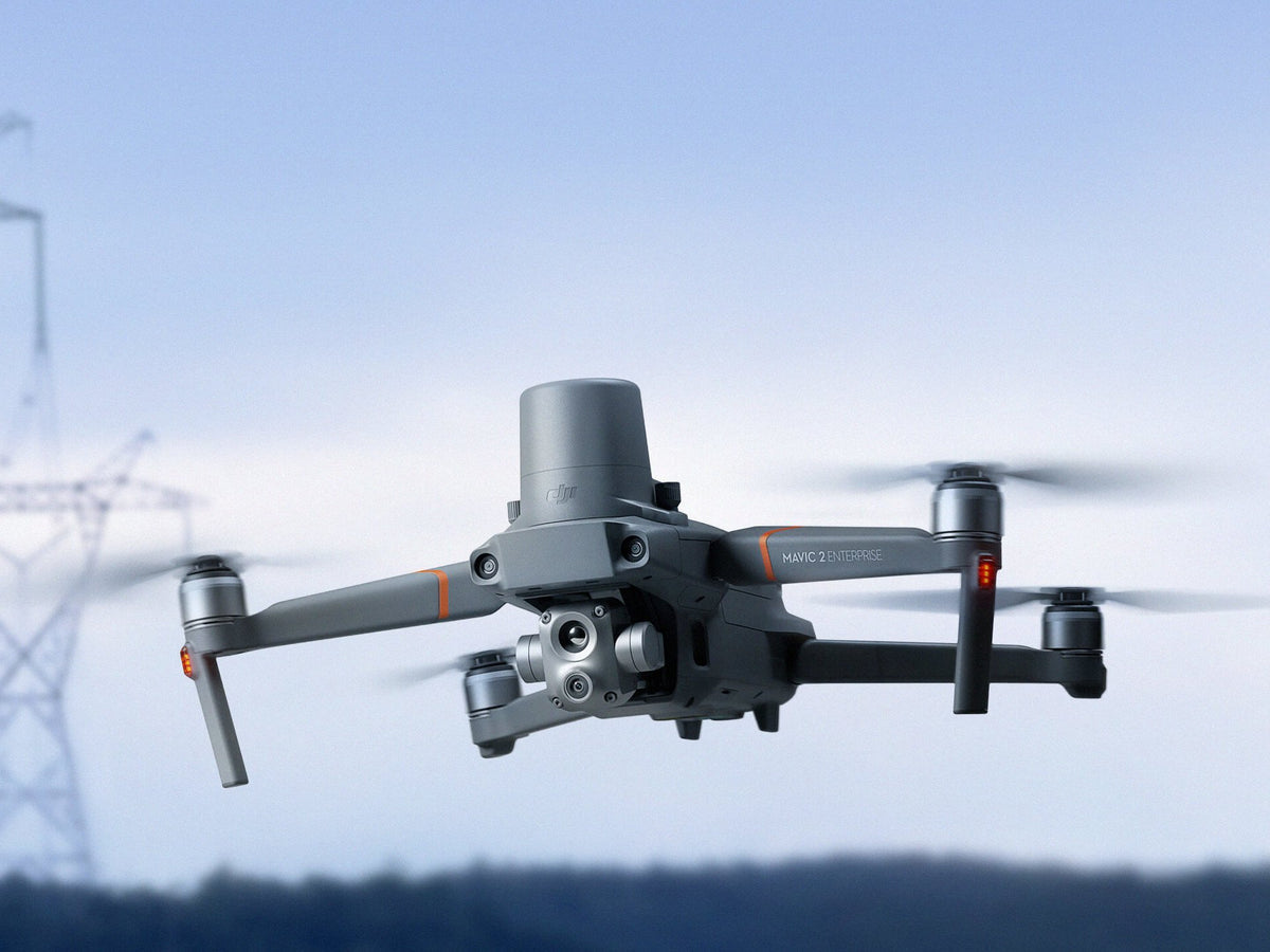 Ein grauer DJI Mavic 2 Enterprise Advanced Multikopter (M2EA) von DJI Enterprise mit Doppelkameras und RTK-Antenne schwebt im Tageslicht, im Hintergrund sind verschwommene Stromleitungen und Bäume zu sehen.