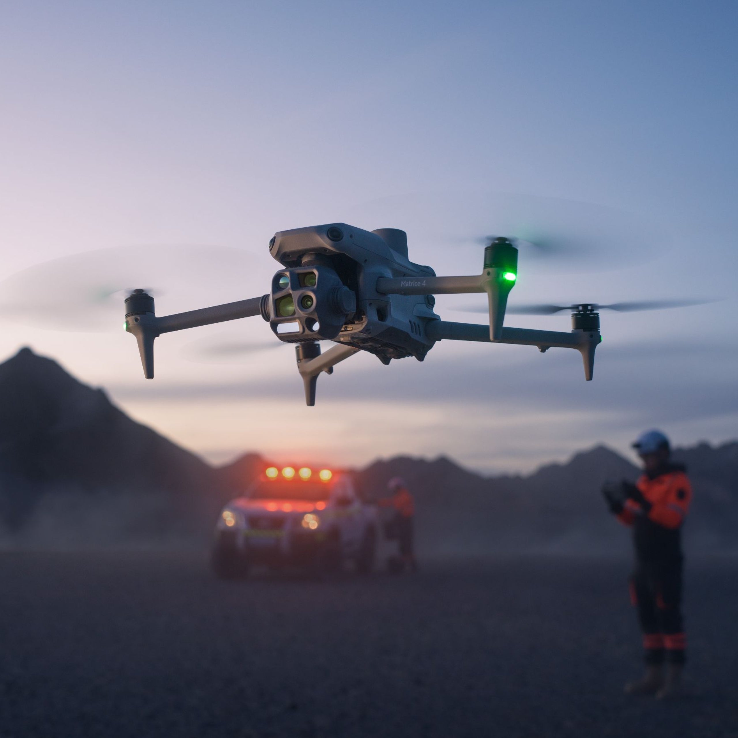 Während die Dämmerung über den Bergen hereinbricht, schwebt eine DJI Matrice 4 Thermal-Drohne von DJI Enterprise mit leuchtenden Lichtern über den Bergen. In der Nähe steht eine Person in einer orangefarbenen Jacke neben einem Fahrzeug mit blinkenden Warnblinklichtern, dessen Silhouette sich vor dem dunklen Himmel abzeichnet.