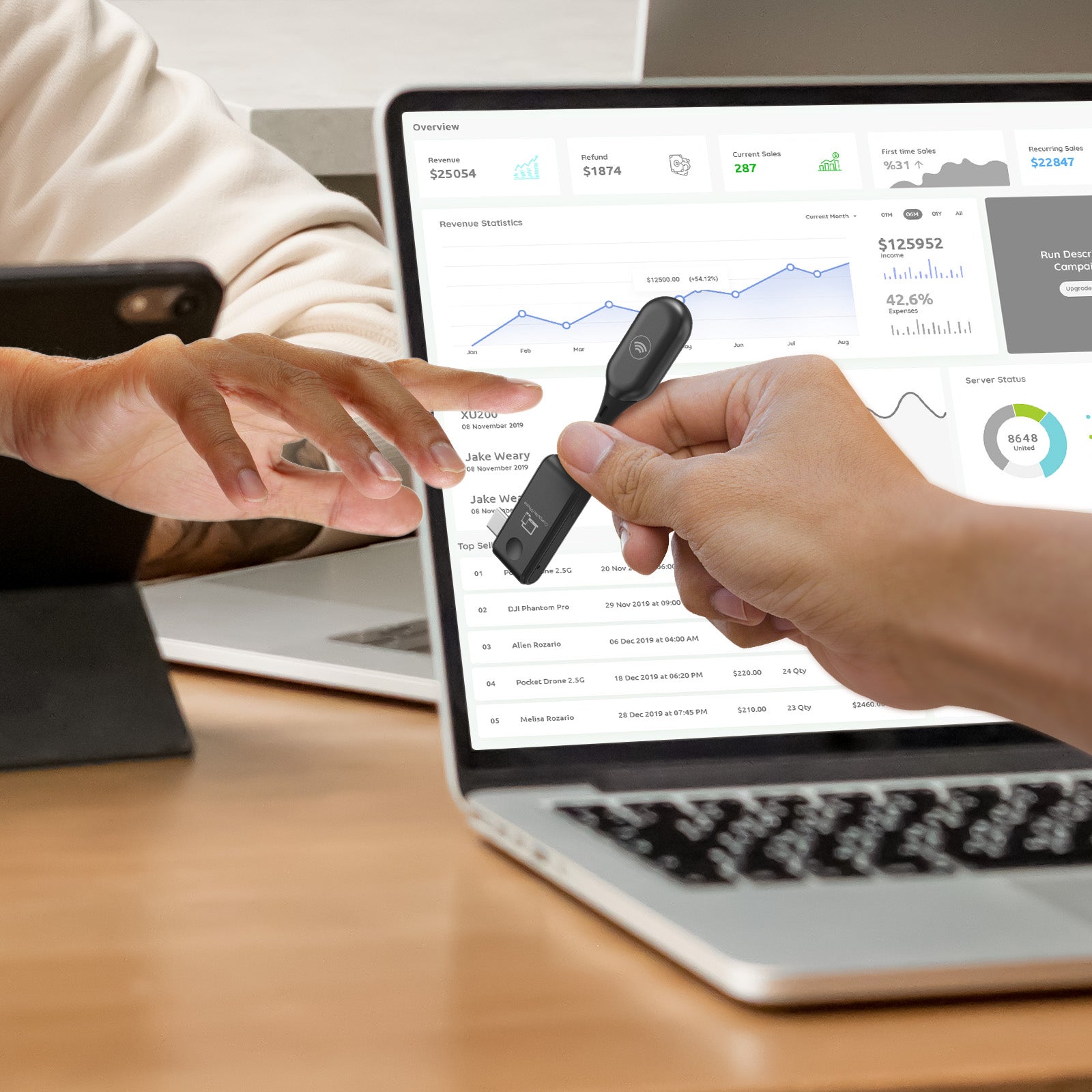 Eine Person übergibt einer anderen Person einen USB-Stick neben einem Laptop mit Grafiken, während im Hintergrund ein MINIX Digitale Bildübertragung - C1, kabelloser USB-C zu HDMI Transmitter, mit einem Tablet auf einem Ständer verbunden ist. Die Umgebung wirkt wie ein Arbeitsplatz.