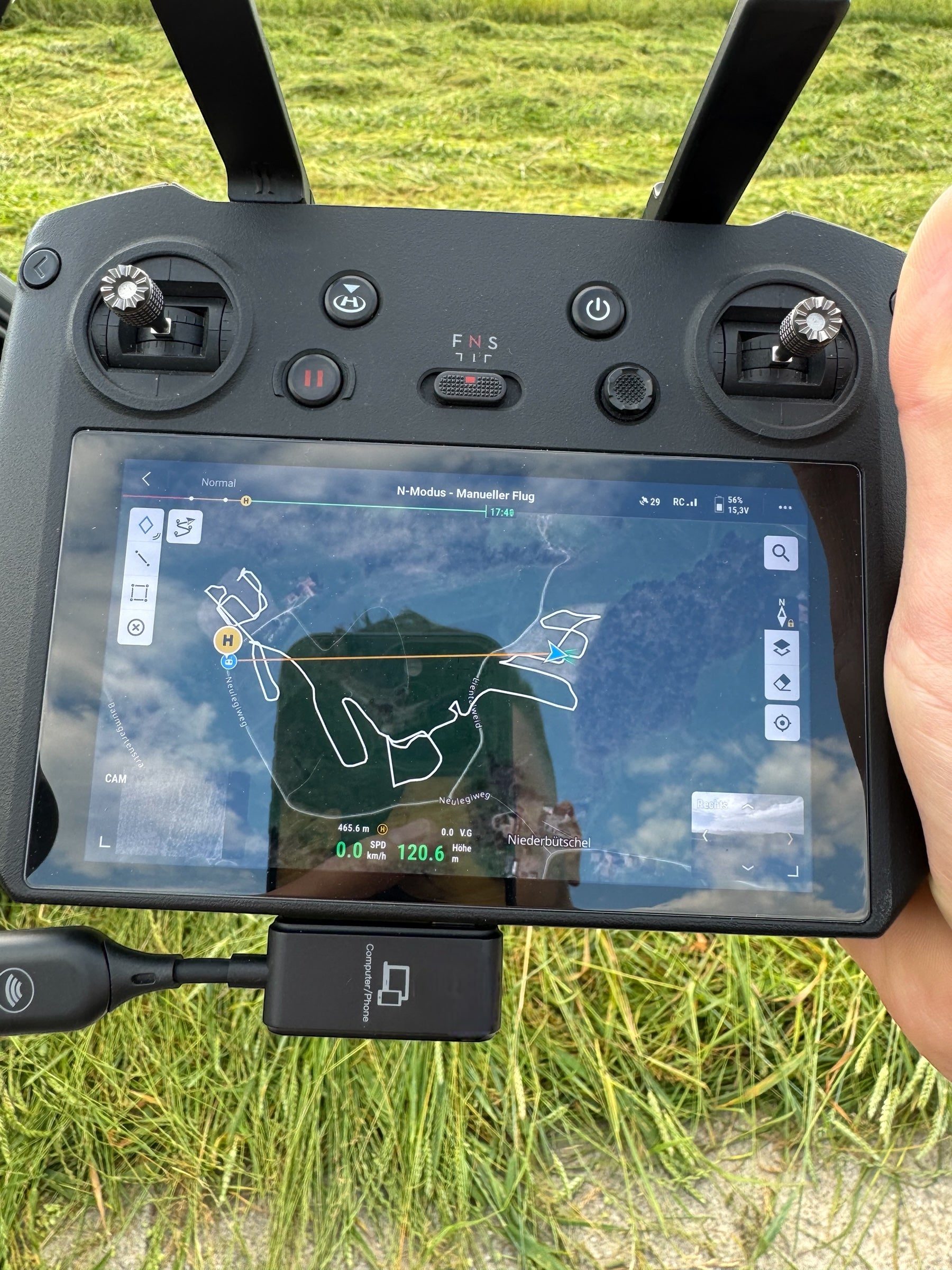 Eine Nahaufnahme zeigt eine Person mit einem Drohnen-Controller, der mit dem kabellosen MINIX C1 USB-C-auf-HDMI-Sender ausgestattet ist. Auf dem Display wird eine Flugkarte angezeigt. Das Display spiegelt teilweise die Wiese wider. Zu sehen sind zwei Steuerknüppel und mehrere Tasten auf dem Controller.