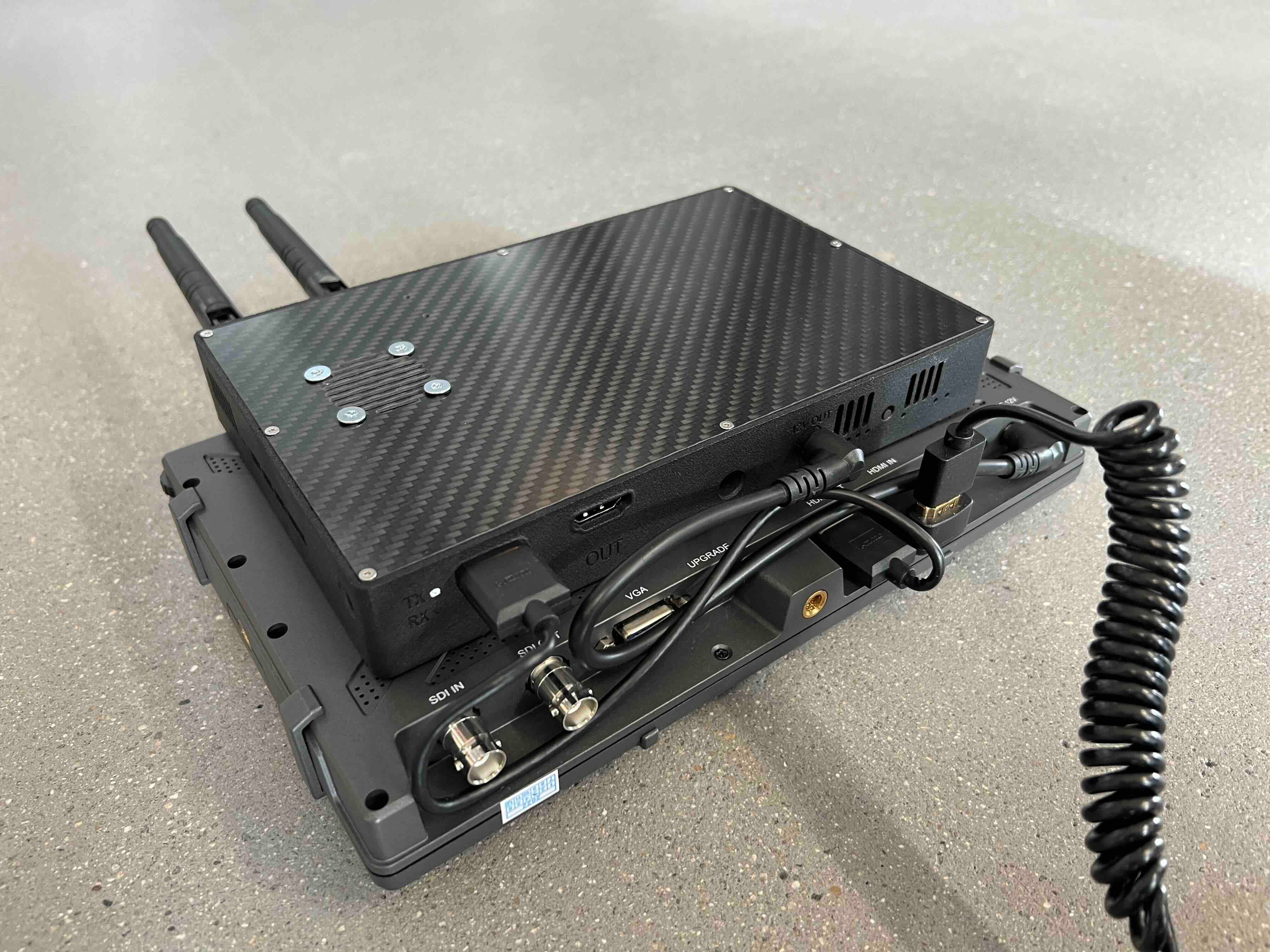 Ein schwarzes elektronisches Gerät mit zwei Antennen und mehreren Anschlüssen ist über Kabel und ein Spiralkabel mit einem anderen Gerät verbunden und liegt auf einer gesprenkelten grauen Oberfläche.