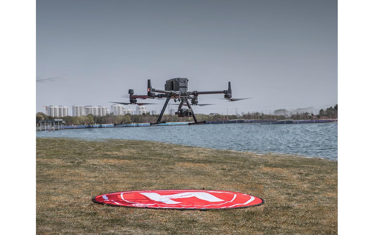 Ein schwarzer DJI-Quadrocopter schwebt über Gras in der Nähe eines roten PGYTECH-Landepads (110 cm) mit wasserdichter Oberfläche am Wasser, mit Gebäuden und Bäumen im Hintergrund unter einem klaren Himmel.