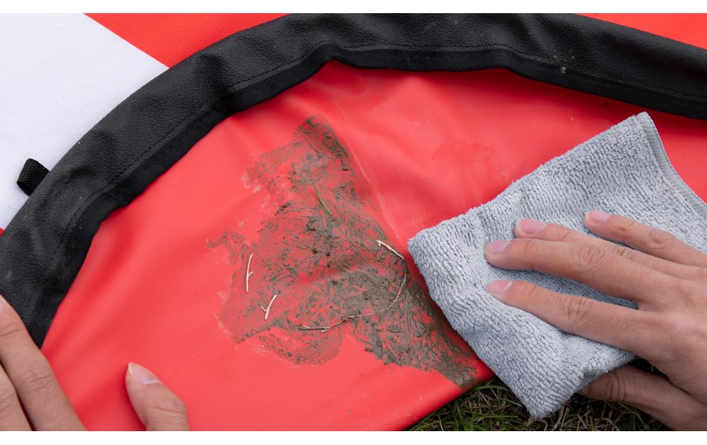 Eine Person verwendet ein graues Tuch, um Schmutz und Grasflecken vom rot-schwarzen PGYTECH Landepad 110 cm zu reinigen, das aus 600D verdicktem Nylon mit einer wasserdichten Oberfläche besteht.
