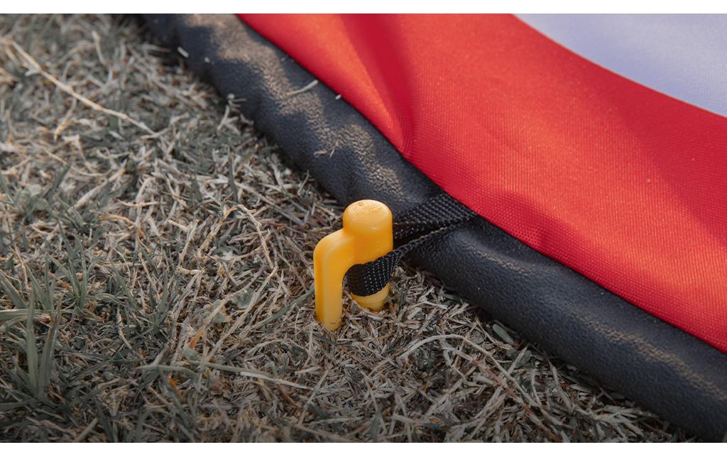 Eine Nahaufnahme eines gelben Kunststoffpfahls, der das PGYTECH Landepad 110 cm aus verdicktem 600D-Nylon und einer wasserdichten Oberfläche auf trockenem Gras befestigt; an der Ecke der rot-weißen Matte ist ein schwarzer Riemen befestigt.