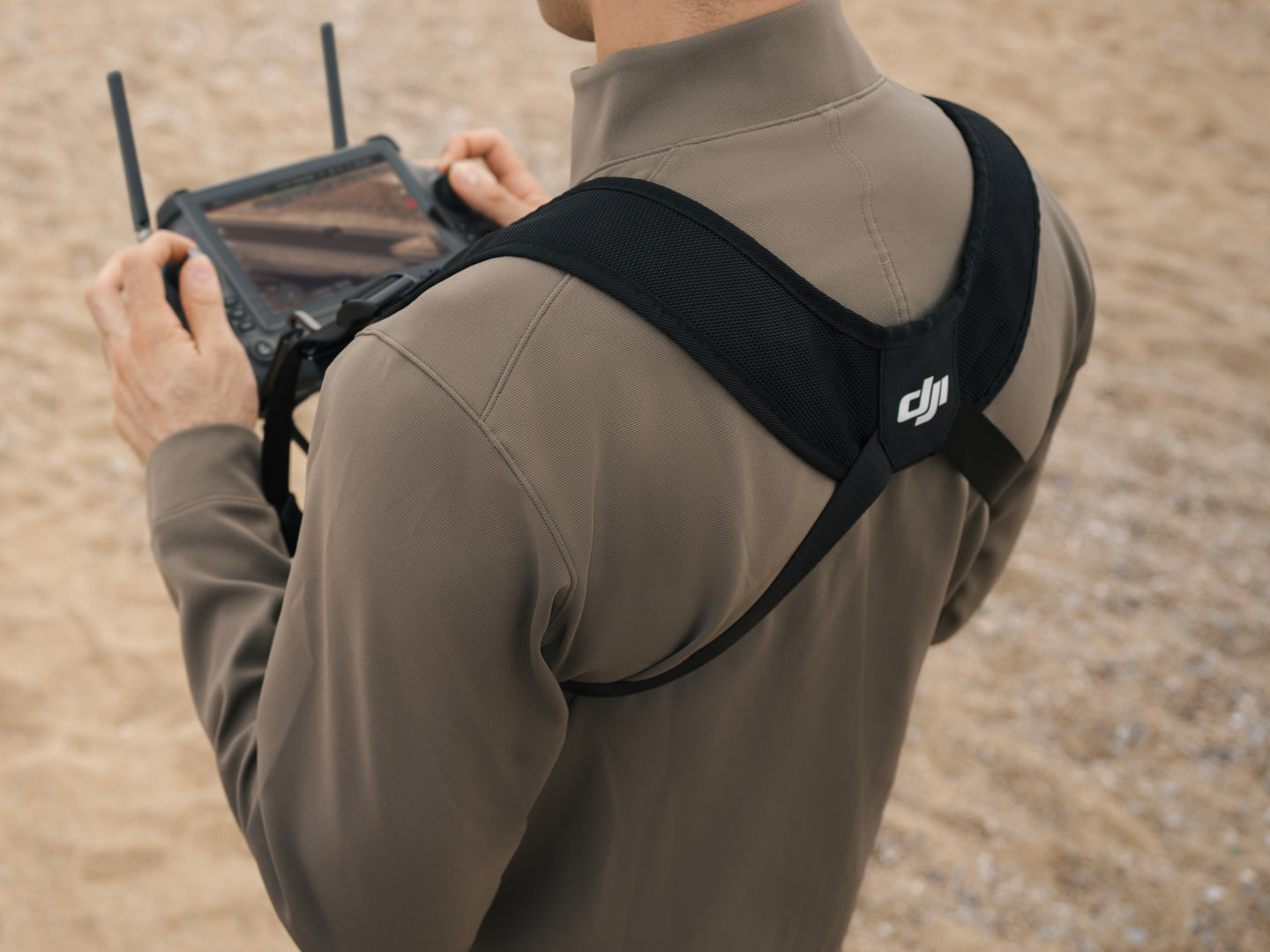 Eine Person in einer beigen Jacke verwendet eine DJI RC Plus-Fernbedienung mit Bildschirm und den DJI Enterprise RC Plus-Gurt und das Taillenstützset (Umhängegurt), während sie auf Sand steht.
