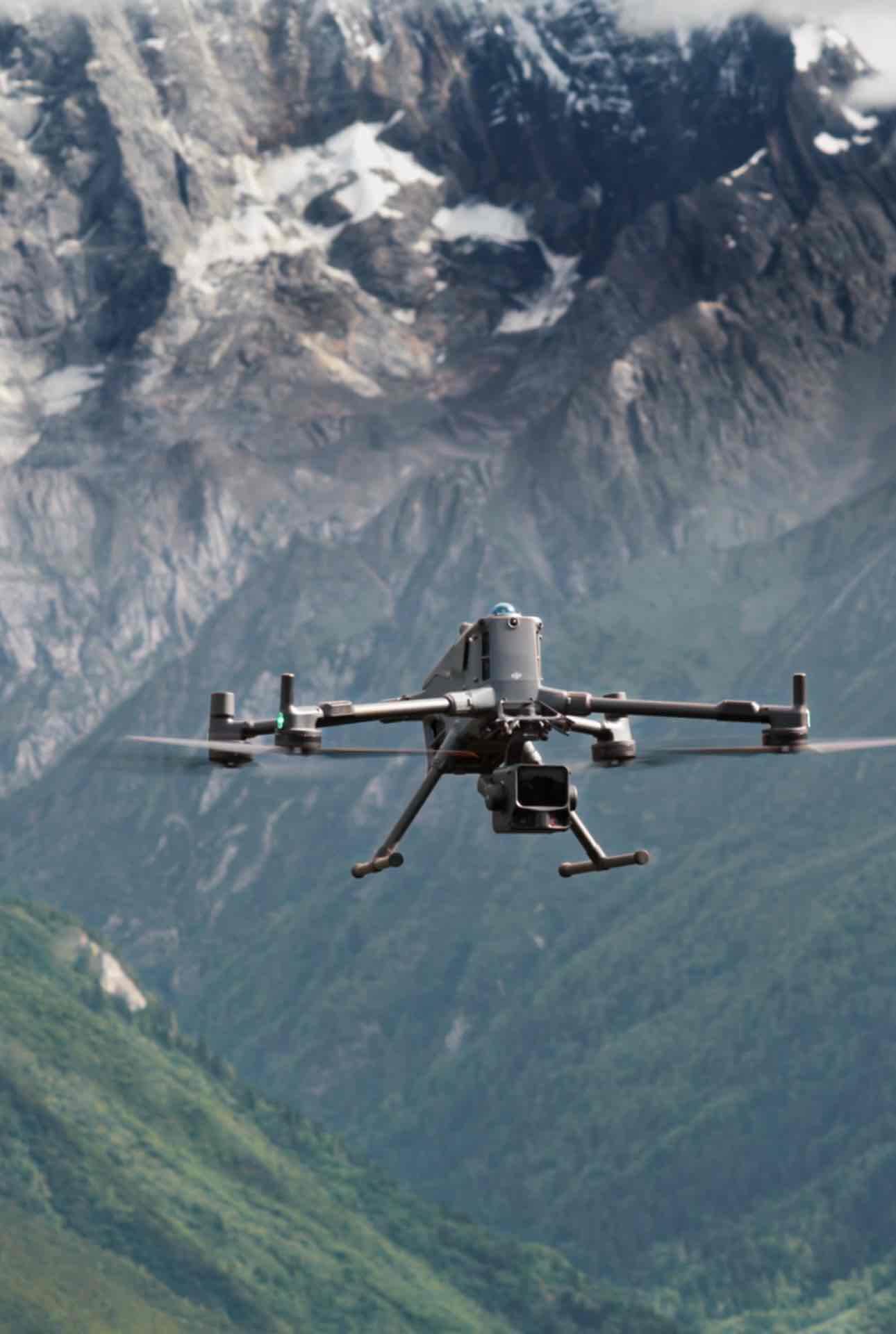 Eine DJI Enterprise Zenmuse L3 Drohne mit hochpräzisem Luft-LiDAR-System und Dual-100MP-RGB-Kameras fliegt über grüne Hügel vor schneebedeckten Bergen und erfasst professionelle Vermessungsdaten unter bewölktem Himmel.