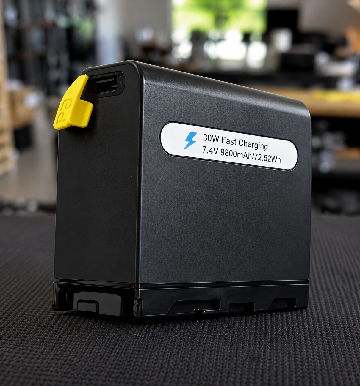 Ein schwarzer CIELOTEC NPF 980 Akku-Monitorakku mit einer gelben Lasche auf der Oberseite, ausgestattet mit integrierter Ladeelektronik, einer Kapazität von 9.800 mAh (7,4 V, 72,52 Wh) sowie PD-30-W-Schnellladefunktion über USB-C und USB-A, liegt auf einer strukturierten Oberfläche in einer sanft unscharf dargestellten Innenraumumgebung – eine vielseitige, mit dem Sony NP-F kompatible Powerbank, die für den längeren Monitorbetrieb konzipiert ist.