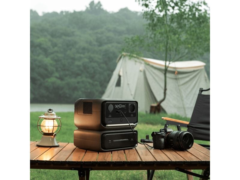 Ein EcoFlow RIVER 3 Max – Powerstation mit 572 Wh / 600 W (X-Boost 1200 W) und LiFePO4-Akku steht auf einem Holztisch auf einem Campingplatz neben einer Kamera und einer Laterne; im Hintergrund sind ein Zelt, ein Stuhl und Bäume in der Nähe eines Flusses oder Sees zu sehen.