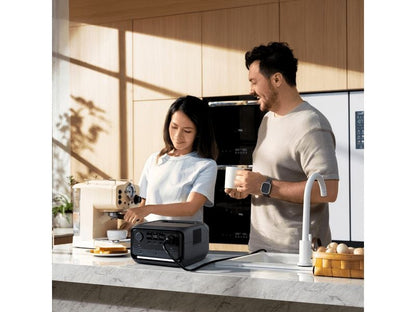 In einer modernen Küche bedient eine Frau eine Espressomaschine, während ein Mann neben ihr lächelt; auf der Arbeitsplatte steht ein EcoFlow RIVER 3 Plus – Powerstation 286 Wh / 600 W (X-Boost 1200 W) LiFePO4 von EcoFlow.