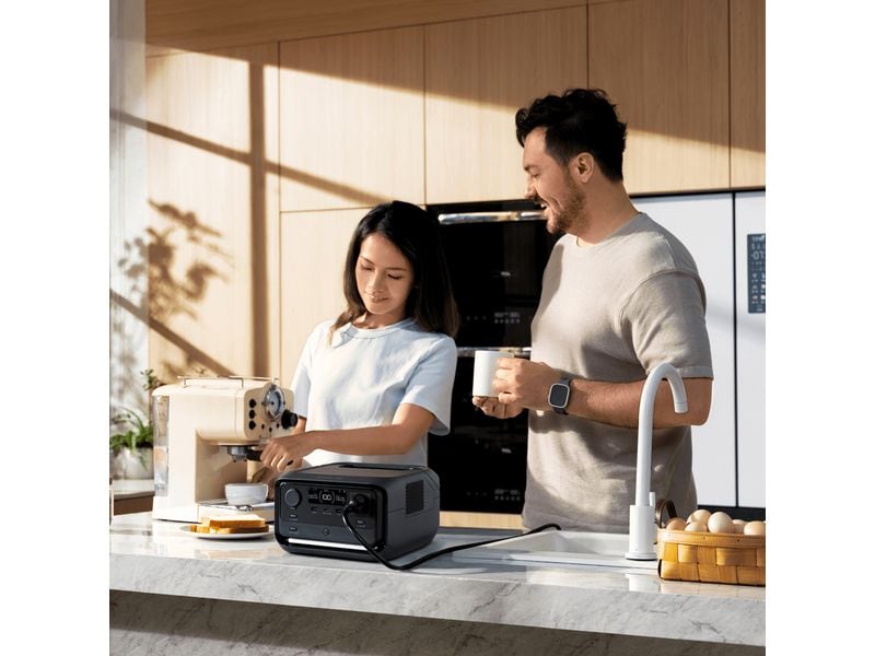 In einer modernen Küche bedient eine Frau eine Espressomaschine, während ein Mann neben ihr lächelt; auf der Arbeitsplatte steht ein EcoFlow RIVER 3 Plus – Powerstation 286 Wh / 600 W (X-Boost 1200 W) LiFePO4 von EcoFlow.