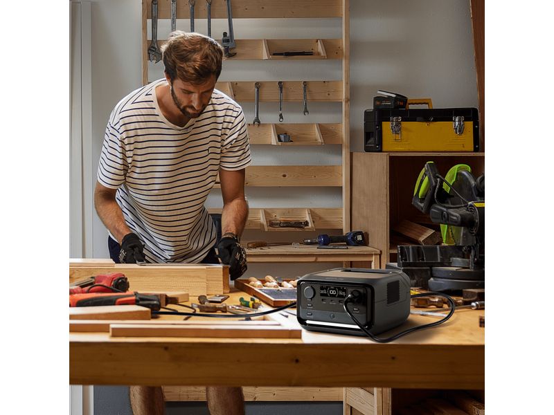 Ein Mann in einem gestreiften Hemd bearbeitet an einer Werkbank Holz. Auf dem Tisch liegen Werkzeuge und eine tragbare Powerstation „EcoFlow RIVER 3 Plus“ von EcoFlow, die über einen 286-Wh-LiFePO4-Akku verfügt und 600 W (X-Boost bis zu 1200 W) liefert. An der Wand hinter ihm sind Regale mit weiteren Werkzeugen angebracht.