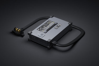 Ein rechteckiges, graues elektronisches Gerät mit einem Griff, einem Netzstecker und Beschriftungen auf der Oberseite – das einem Zubehörteil für die DJI Delivery FlyCart 100 Schwerlast-Transportdrohne mit Fallschirm (bis zu 100 kg Nutzlast, 12 km Reichweite) ähnelt – liegt auf einer dunklen Oberfläche.