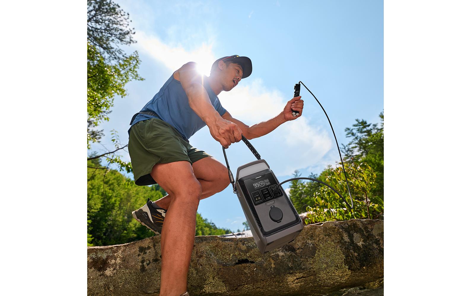 Eine Person in Outdoor-Kleidung kniet auf einem Baumstamm inmitten sonnenbeschienener Bäume und hält die EcoFlow TRAIL Plus 300 DC in der Hand – Powerstation 288 Wh / 300 W (USB-C 140 W) mit Campinglampe von EcoFlow und angeschlossenen Kabeln in der Hand – dank seiner zuverlässigen Kapazität von 288 Wh ein idealer Begleiter für das Powerstation-Camping.