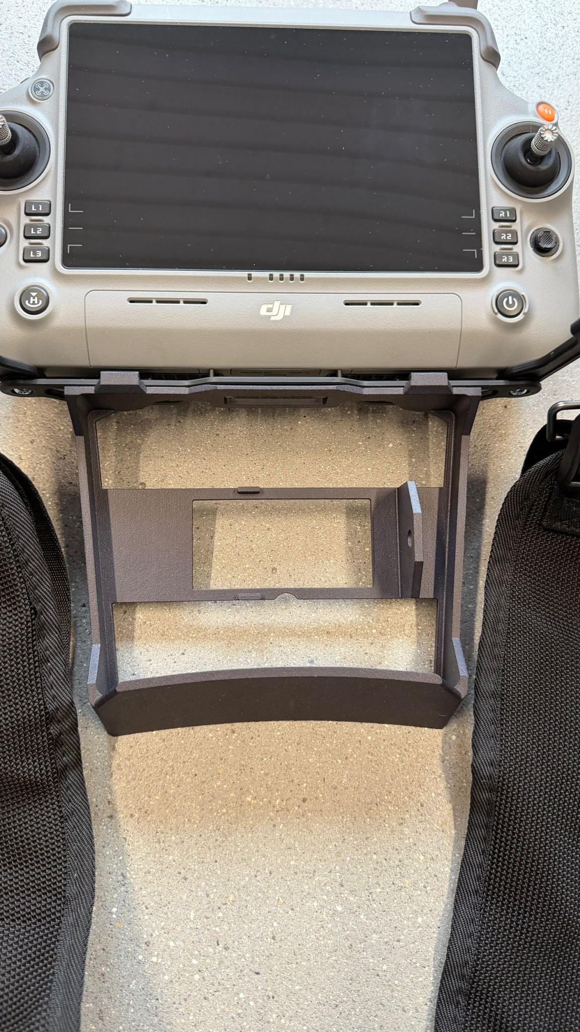 Ein DJI-Drohnen-Controller, ausgestattet mit einer schwarzen CIELOTEC RC Plus 2-Distanzhalterung – SDR- & Lautsprecher-Version (3D-Druck PA12), die für die Verwendung mit dem DJI RC Plus 2-Sendertragegurt (M4E, M4T) ist darunter angebracht und ruht auf einer gesprenkelten Oberfläche, wobei auf beiden Seiten schwarze Stoffriemen und ein DJI-Sendertragegurt zu sehen sind.